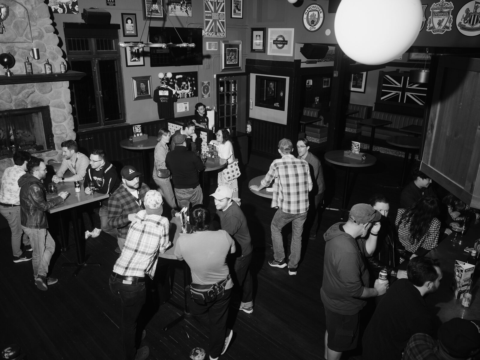 High view over the busy pub floor, attendees clustered around high-tops beside a stone fireplace, Union Jack art and framed football photos on the walls.