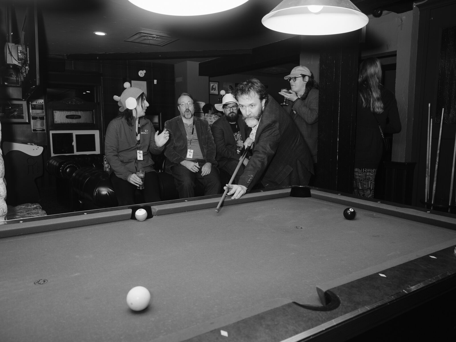 A bearded attendee in a blazer grins at the camera mid-shot, while attendees in a knit animal hat and a cowboy hat watch from the leather couch.