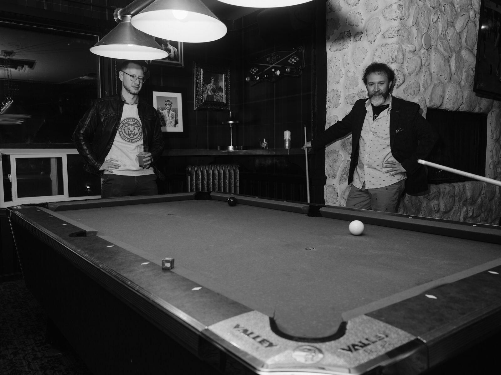 Two attendees stand at opposite ends of the pool table, one in a leather jacket holding a drink, the other leaning on their cue by the stone wall.