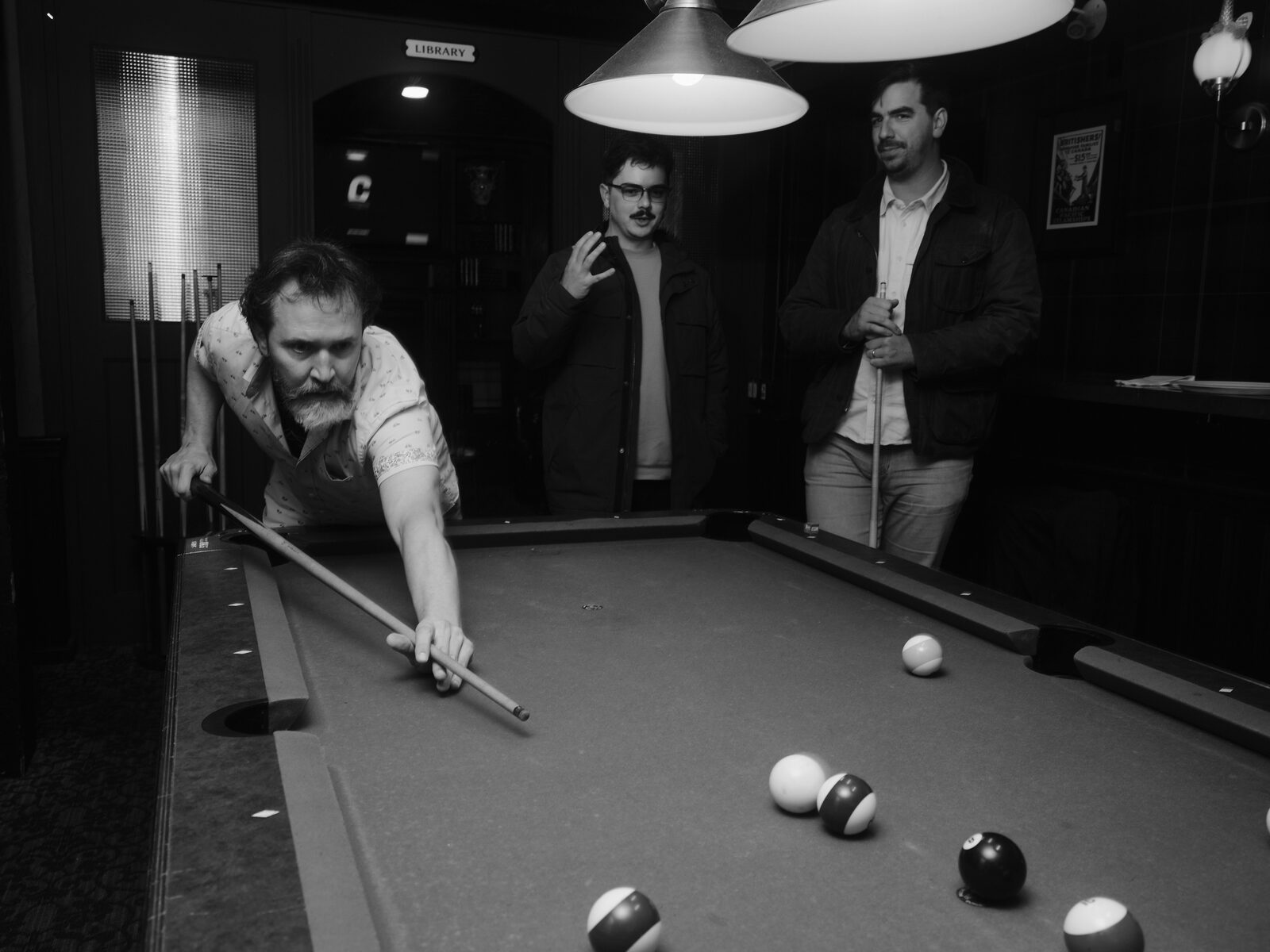 A bearded player in a short-sleeved print shirt stretches out a long shot as two attendees watch, one mid-gesture, near the pub's "LIBRARY" doorway.