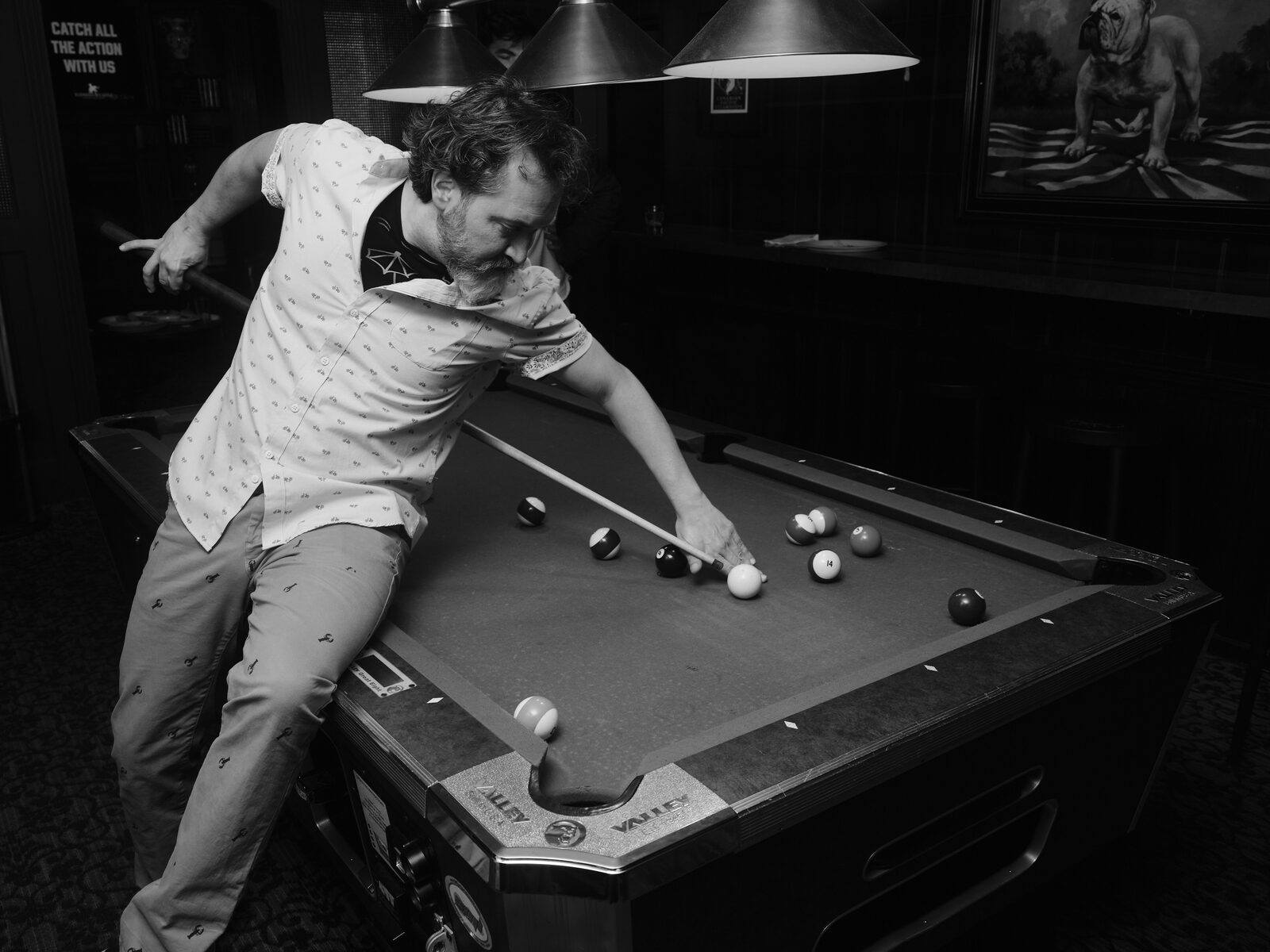 A bearded player in a print shirt perches one leg on the rail and reaches behind their back for a trick shot, the bulldog painting looming behind.