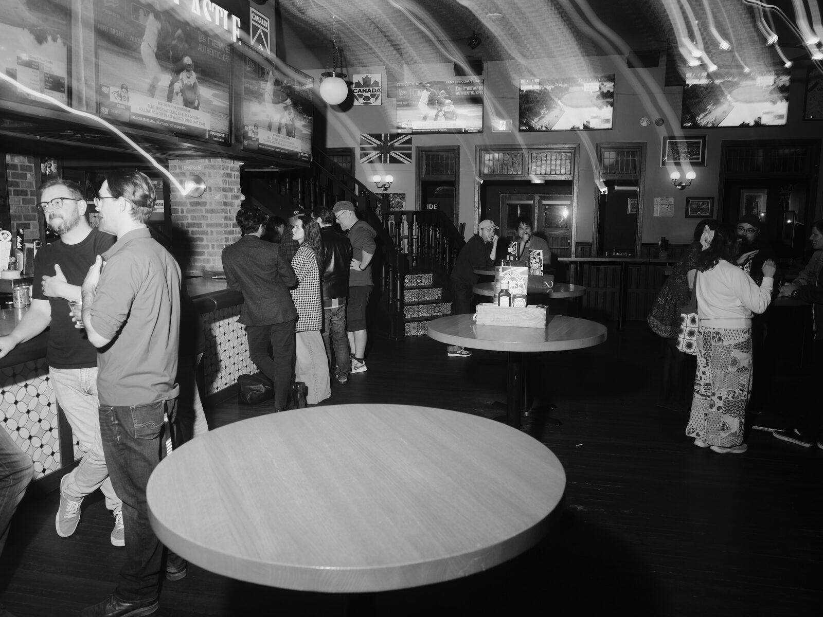 A long-exposure shot of the pub floor, light trails streaking across the frame over attendees at the bar and high-tops beneath baseball on the TVs.
