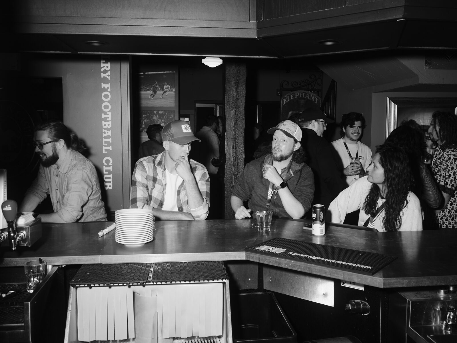 Attendees lean on the bar across from the camera, one in a ball cap and flannel resting their chin on a hand beside a stack of plates.