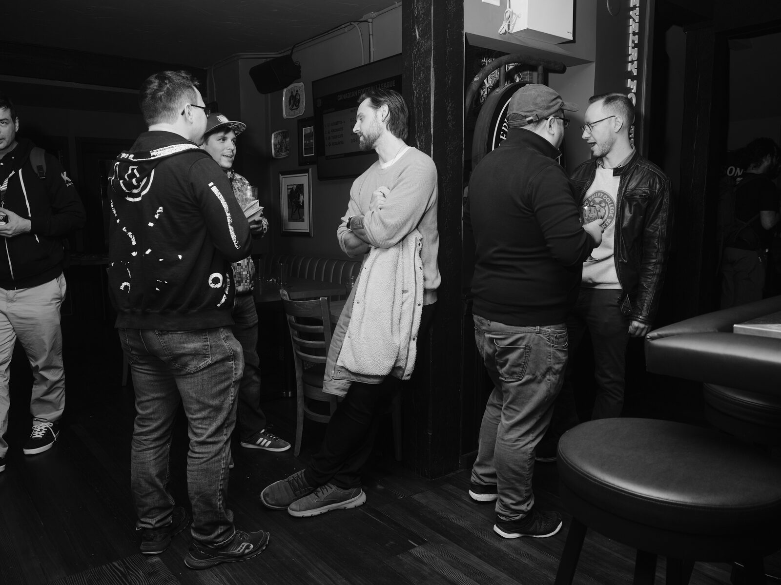 Five attendees stand in a loose circle chatting near a dartboard in a side room, one holding a fleece jacket over their arm.