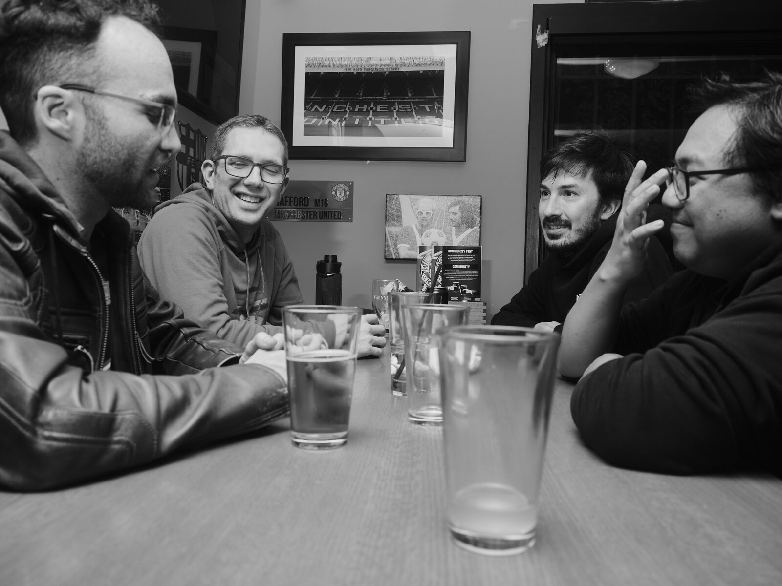 Four attendees in glasses laugh and talk around a table of pint glasses, framed Manchester United photos on the wall behind.