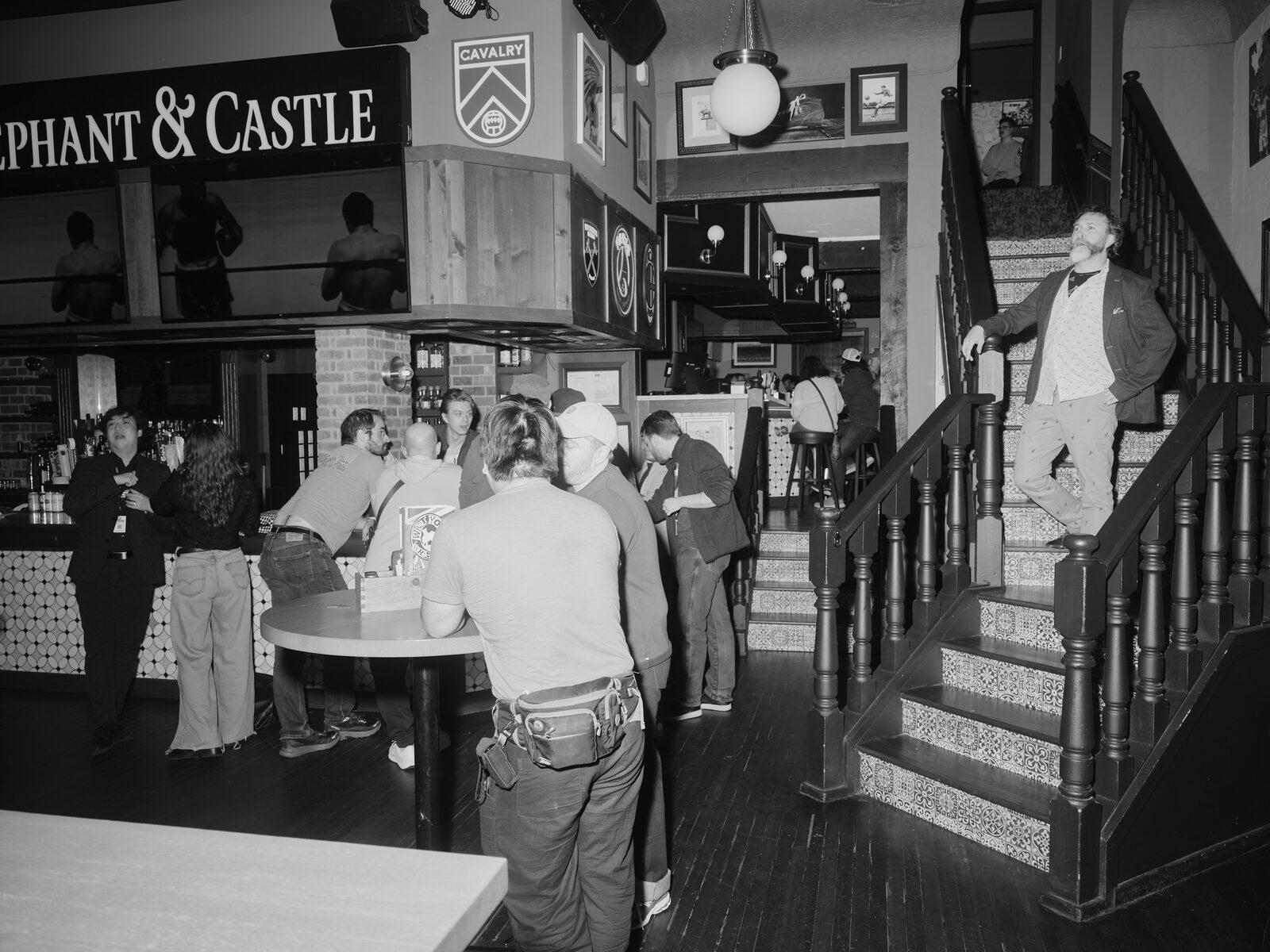 An attendee in a blazer strikes a pose on the tiled staircase under the Elephant & Castle sign and Cavalry FC crest, attendees mingling at the bar below.