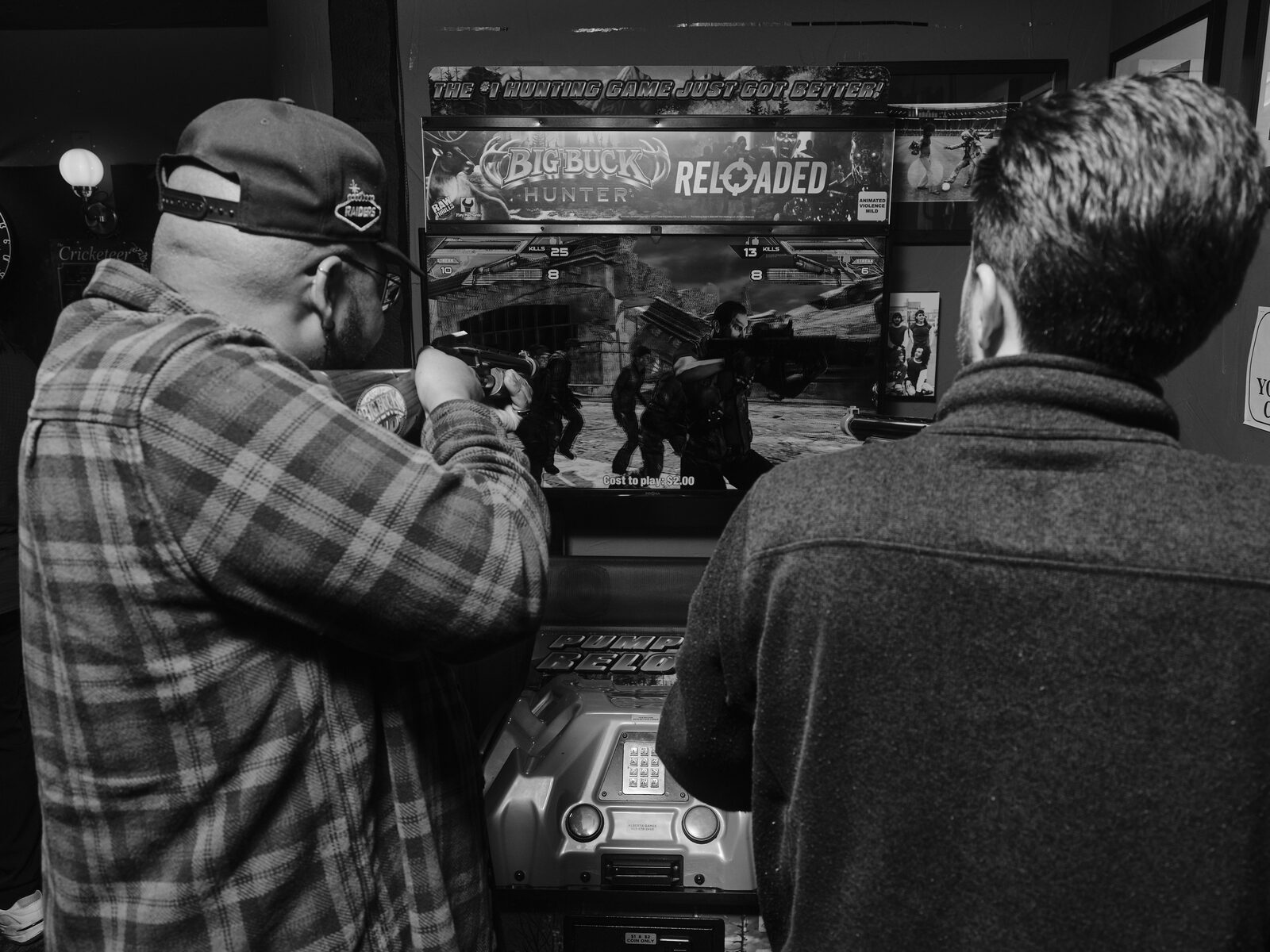 An attendee in a flannel and backwards cap aims a plastic rifle at the Big Buck Hunter Reloaded arcade cabinet as a friend watches.