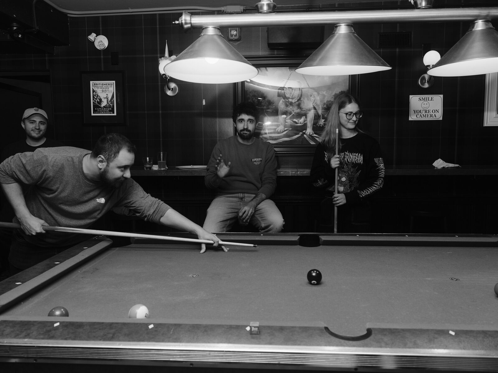 A player in a grey tee lines up a shot on the pool table as three attendees look on, one gesturing, beneath the bulldog painting and hanging lamps.