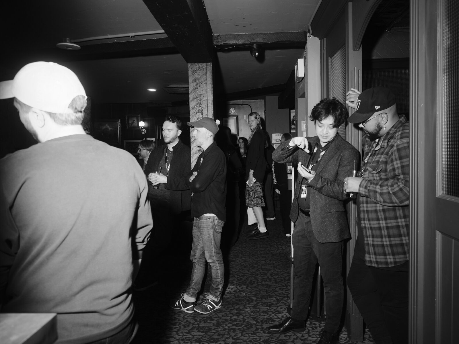 Attendees chat along a narrow carpeted hallway of the pub, one checking their phone in a doorway while another lifts a hand to their cap.