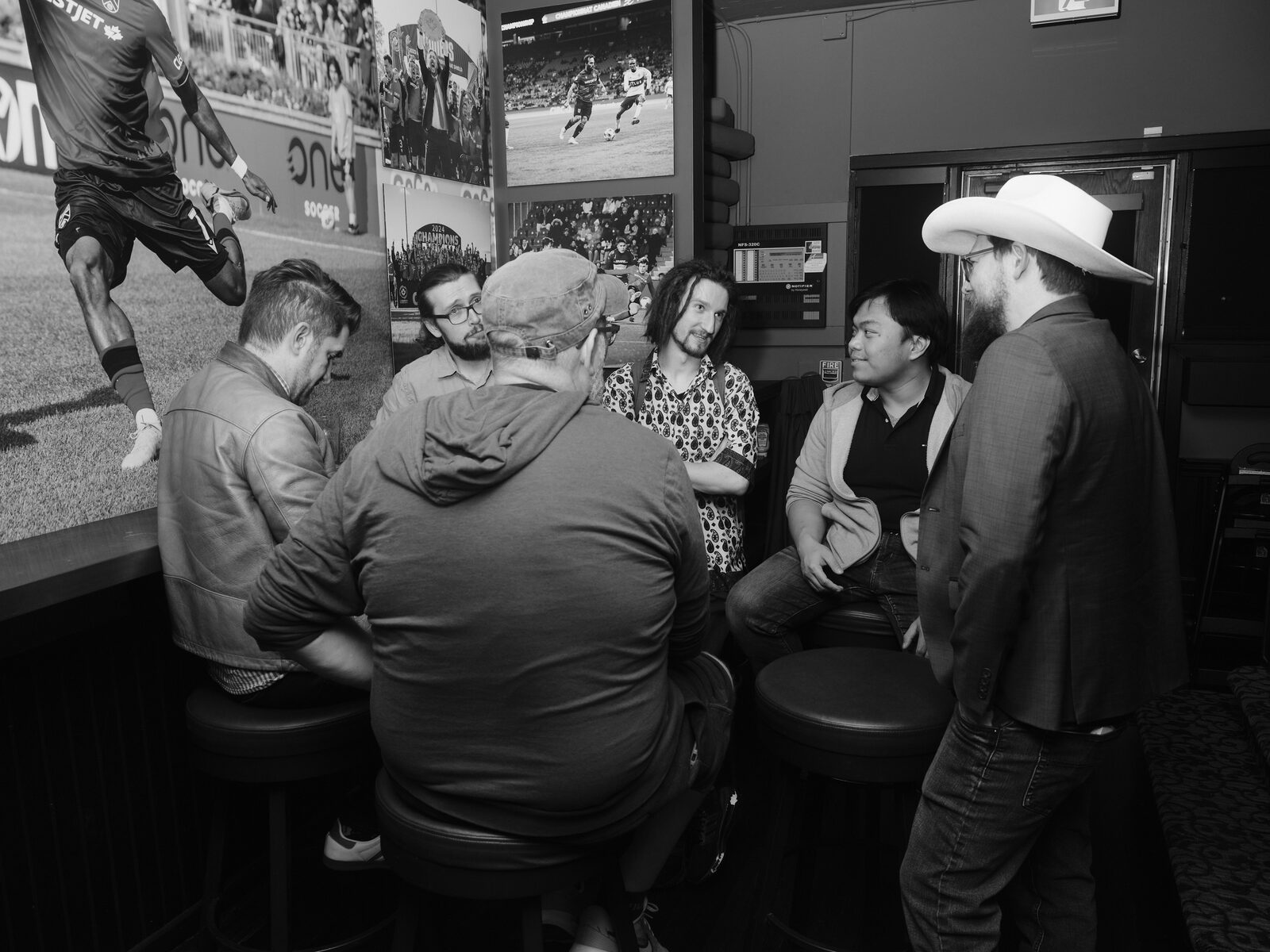 Five attendees gather around bar stools beneath a big football mural, one in a white cowboy hat and blazer standing over the conversation.