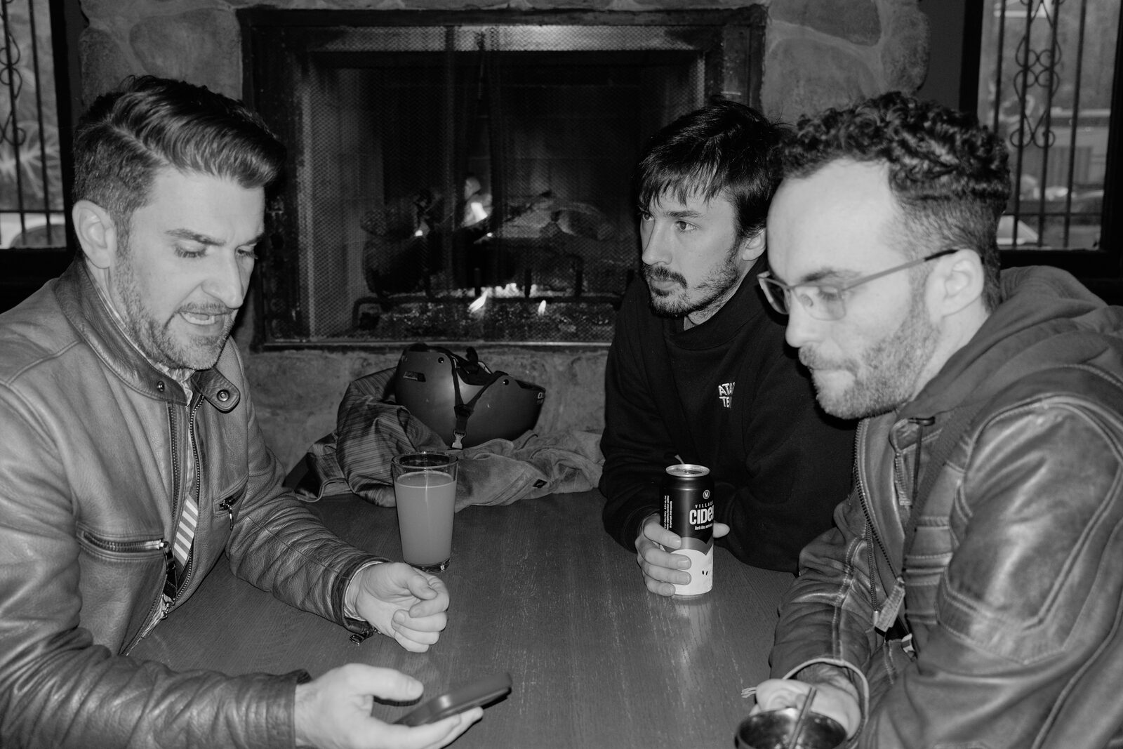 Three attendees in leather jackets lean over a table in front of a lit fireplace, one checking a phone, another holding a can of cider.