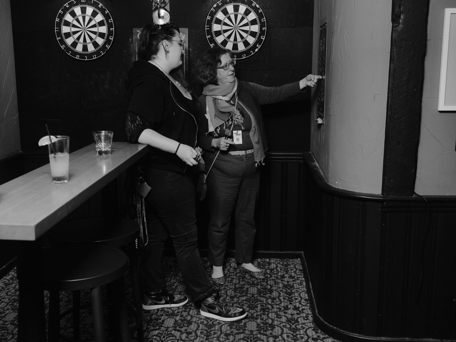 Two attendees play darts at a pair of boards on the back wall, one reaching up to the scoreboard, drinks on the ledge beside them.