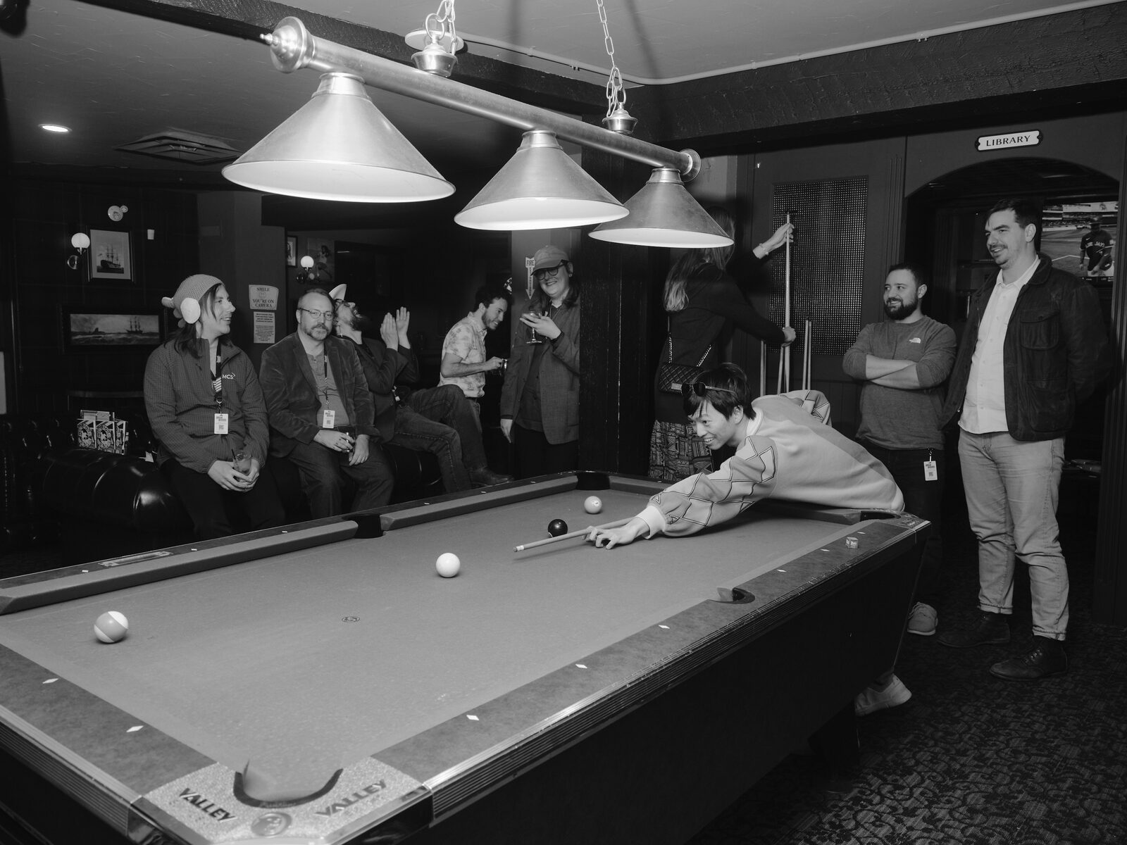 An attendee in a patterned cardigan stretches across the pool table for a shot as a row of attendees on the couch cheers and laughs under the "LIBRARY" sign.