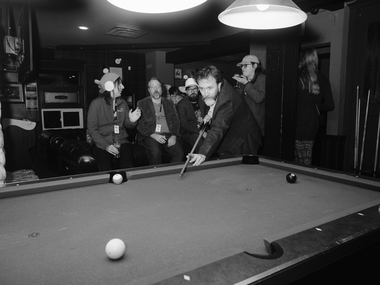 A bearded attendee in a blazer grins at the camera mid-shot, while attendees in a knit animal hat and a cowboy hat watch from the leather couch.