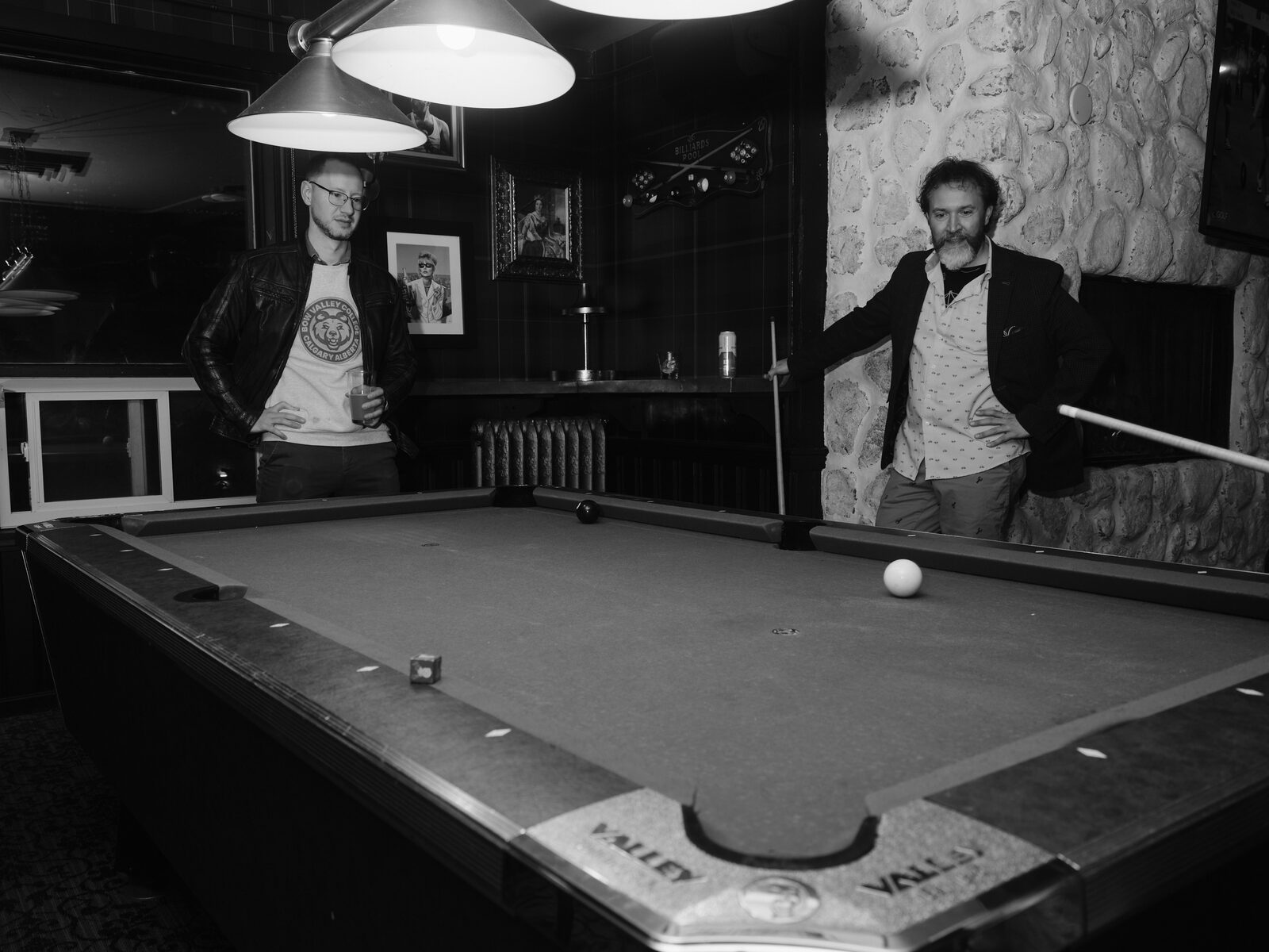 Two attendees stand at opposite ends of the pool table, one in a leather jacket holding a drink, the other leaning on their cue by the stone wall.