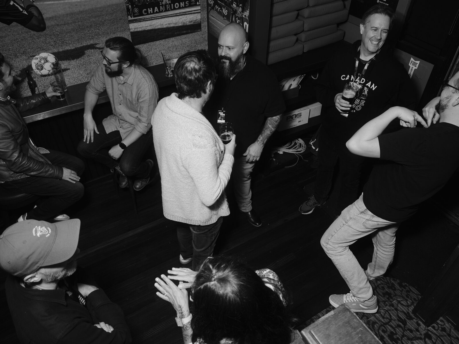 High view of a lively corner of the pub, one attendee mid-dance move while others laugh and chat with pints, a football "CHAMPIONS" mural on the wall.