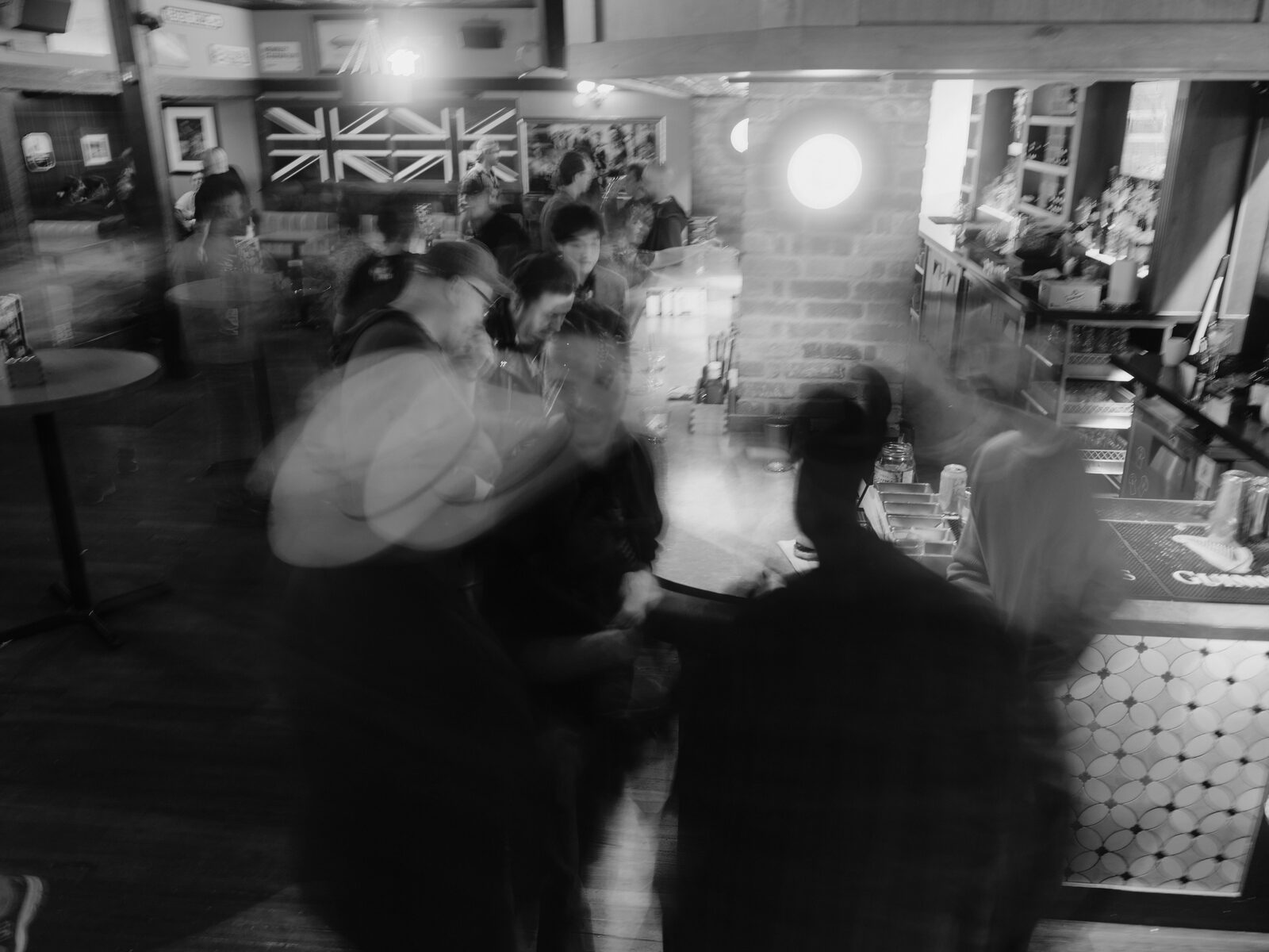 A long exposure turns the crowd along the bar into a blur of motion, a Union Jack and a bright round lamp on the wall behind.