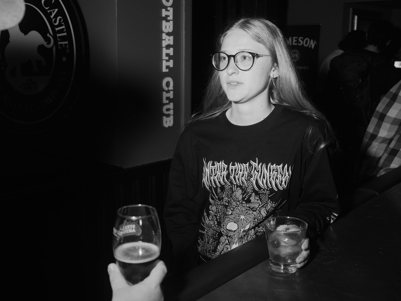 An attendee in round glasses and a metal-band long-sleeve stands at the bar beside the Elephant & Castle football club crest, a pint held out toward them from out of frame.