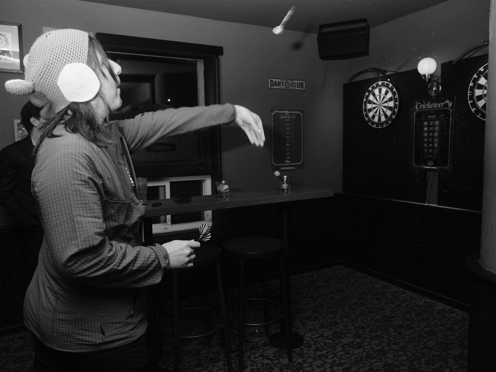 An attendee in a knit animal hat follows through on a dart throw, the dart a blur in the air, heading for the boards beneath the "DART CLUB" sign.