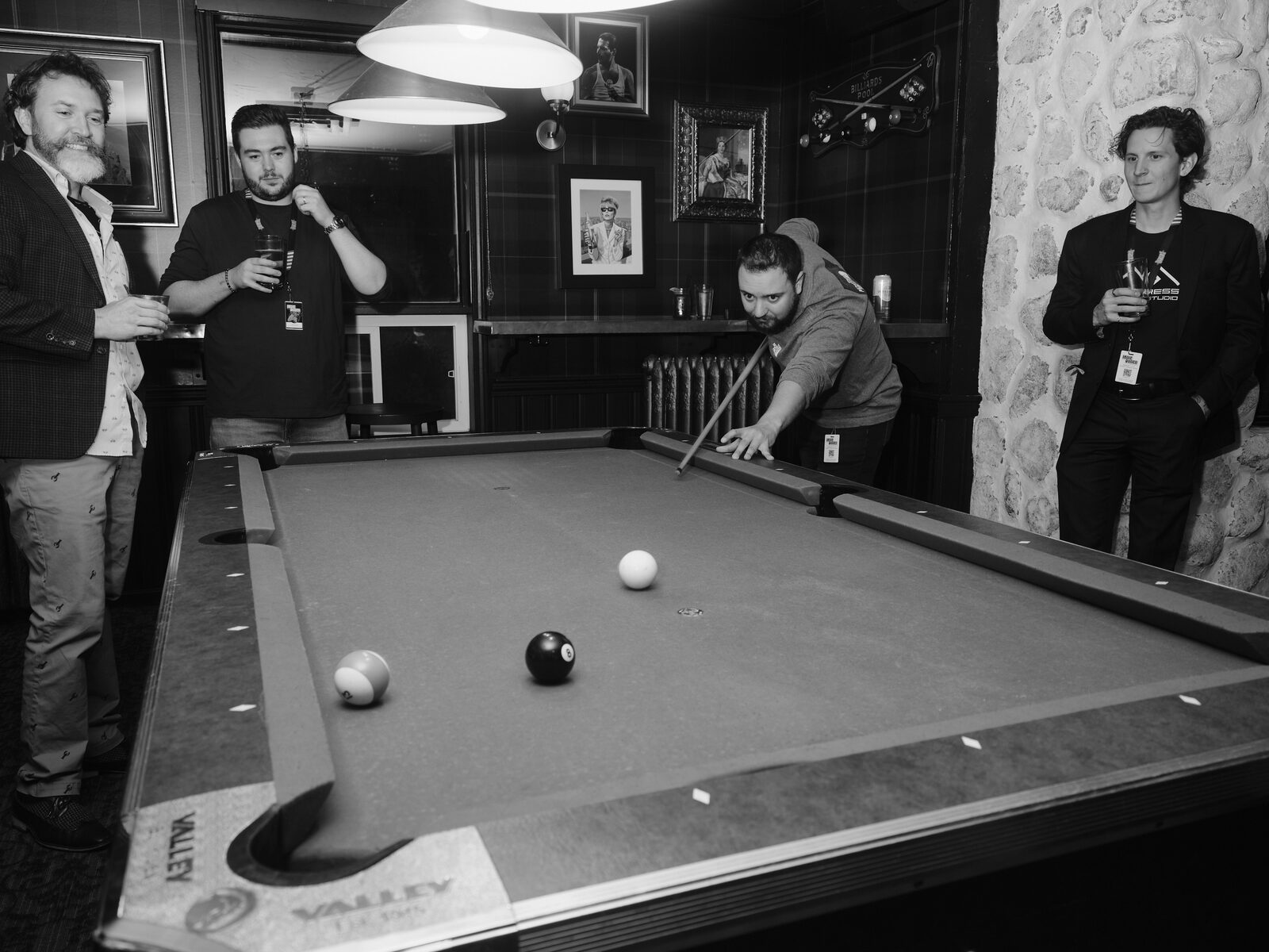 A player in a hoodie sinks low for a shot on the Valley pool table while three attendees holding drinks watch from around the room.