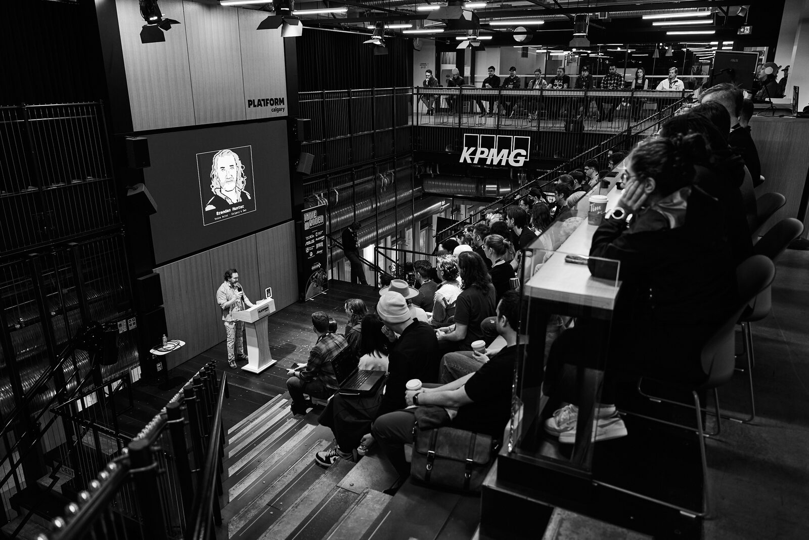 Looking down into the Platform Calgary atrium from the balcony: a speaker at the lit podium, their portrait on the big screen, attendees filling the bleacher steps under the KPMG sign.
