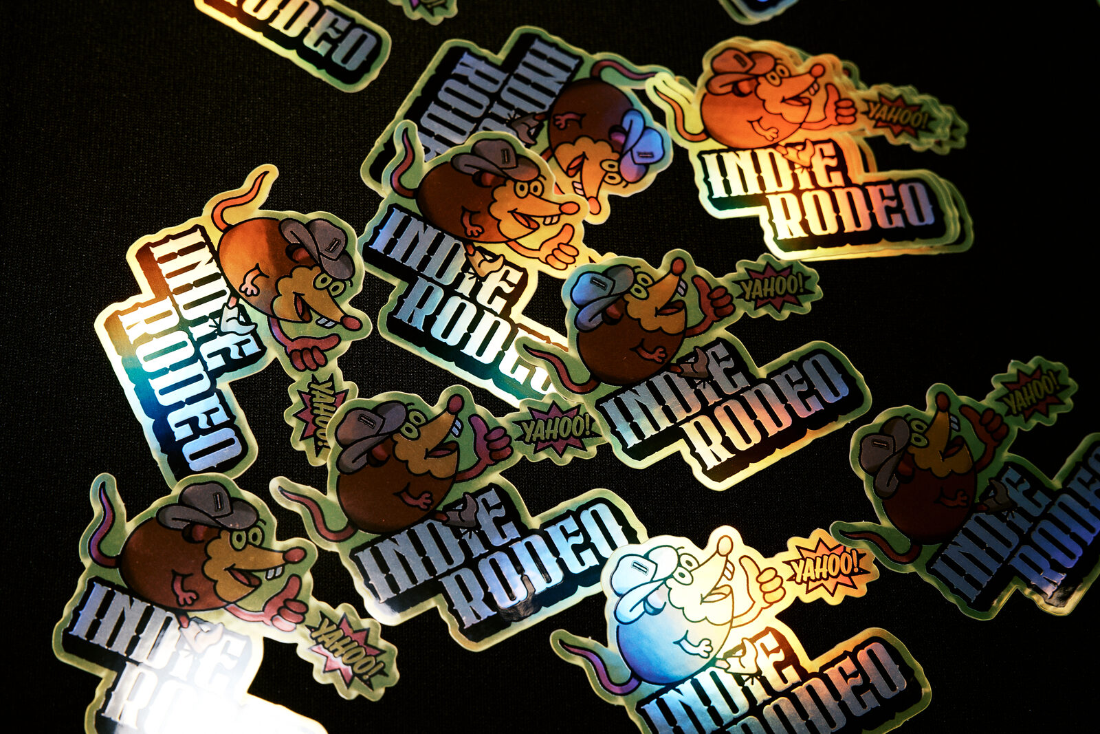 A scattered pile of holographic Indie Rodeo stickers on a black tablecloth — the grinning rat mascot and the wordmark, catching rainbow light.