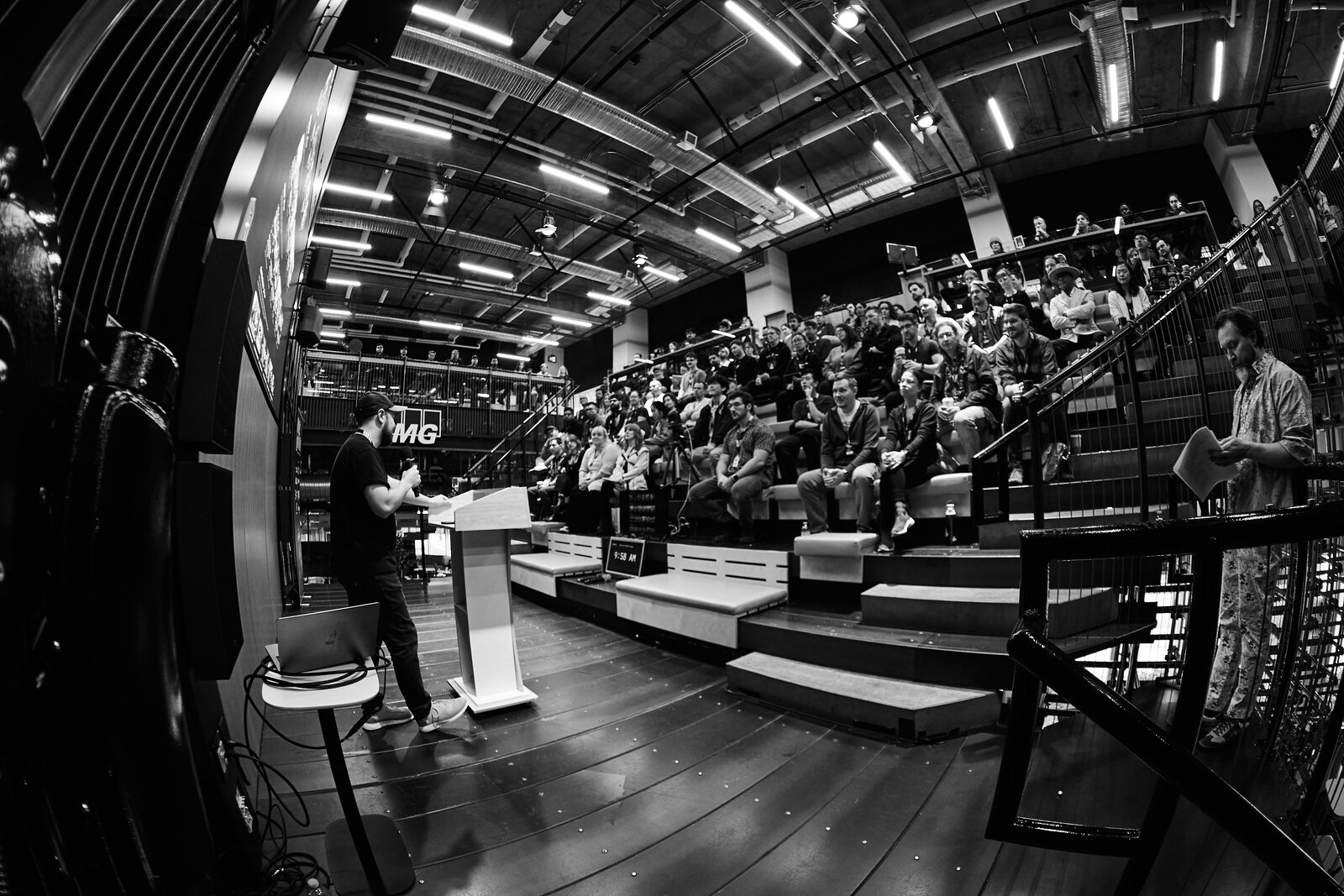 Fisheye view from beside the stage: a speaker at the podium facing a packed house of attendees on the bleachers.