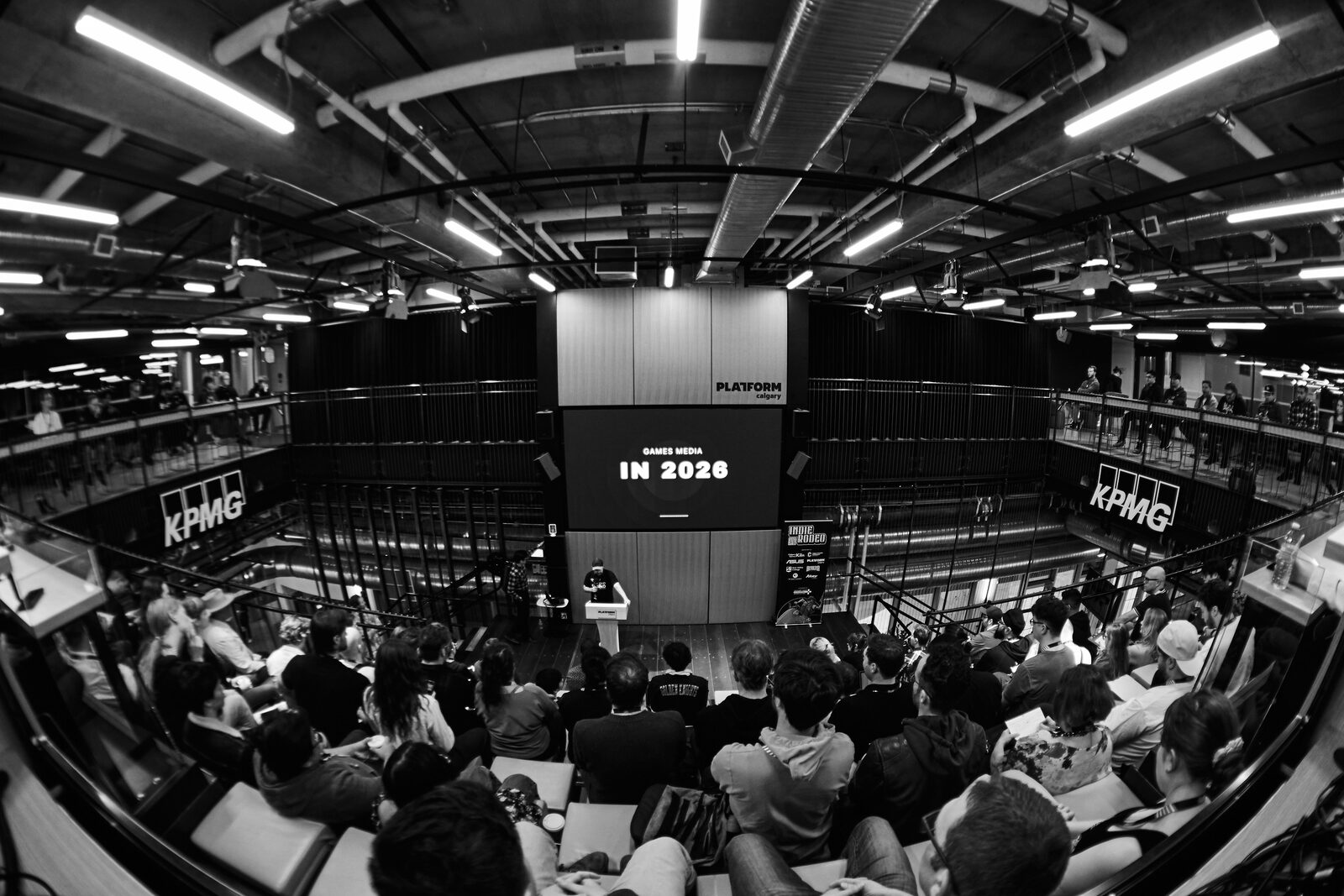 Fisheye view from the back of the room, the full atrium in frame, the stage screen reading "IN 2026" over the seated crowd.