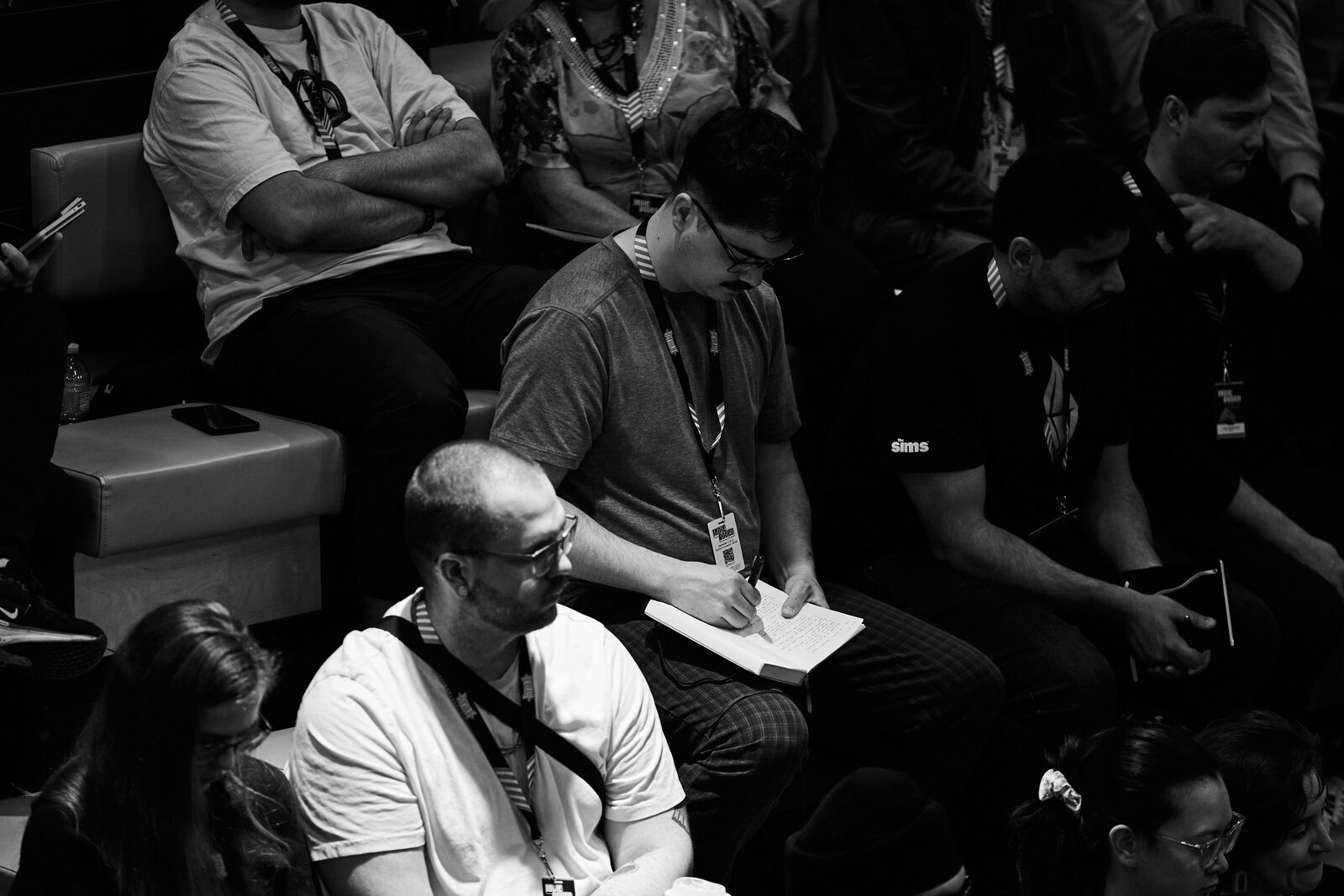 Attendees on the bleachers during a talk; one of them is taking notes in a paper notebook.