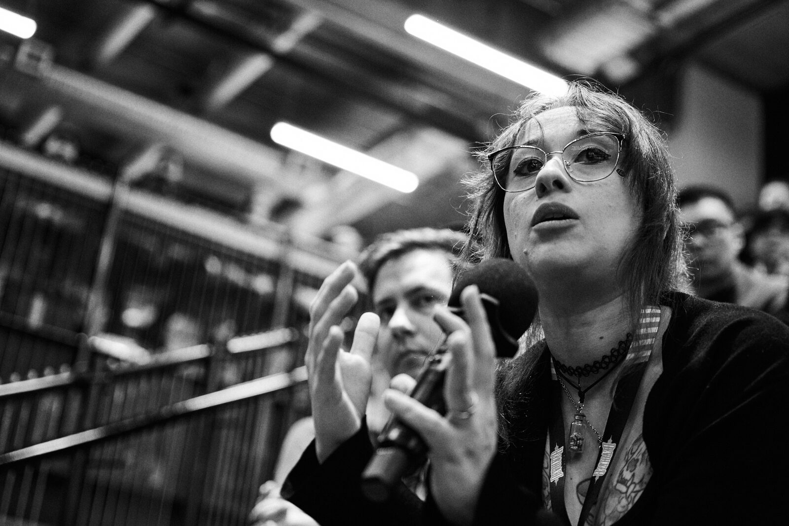 An attendee with glasses gestures as she asks a question, mic in hand, a second attendee listening beside her.