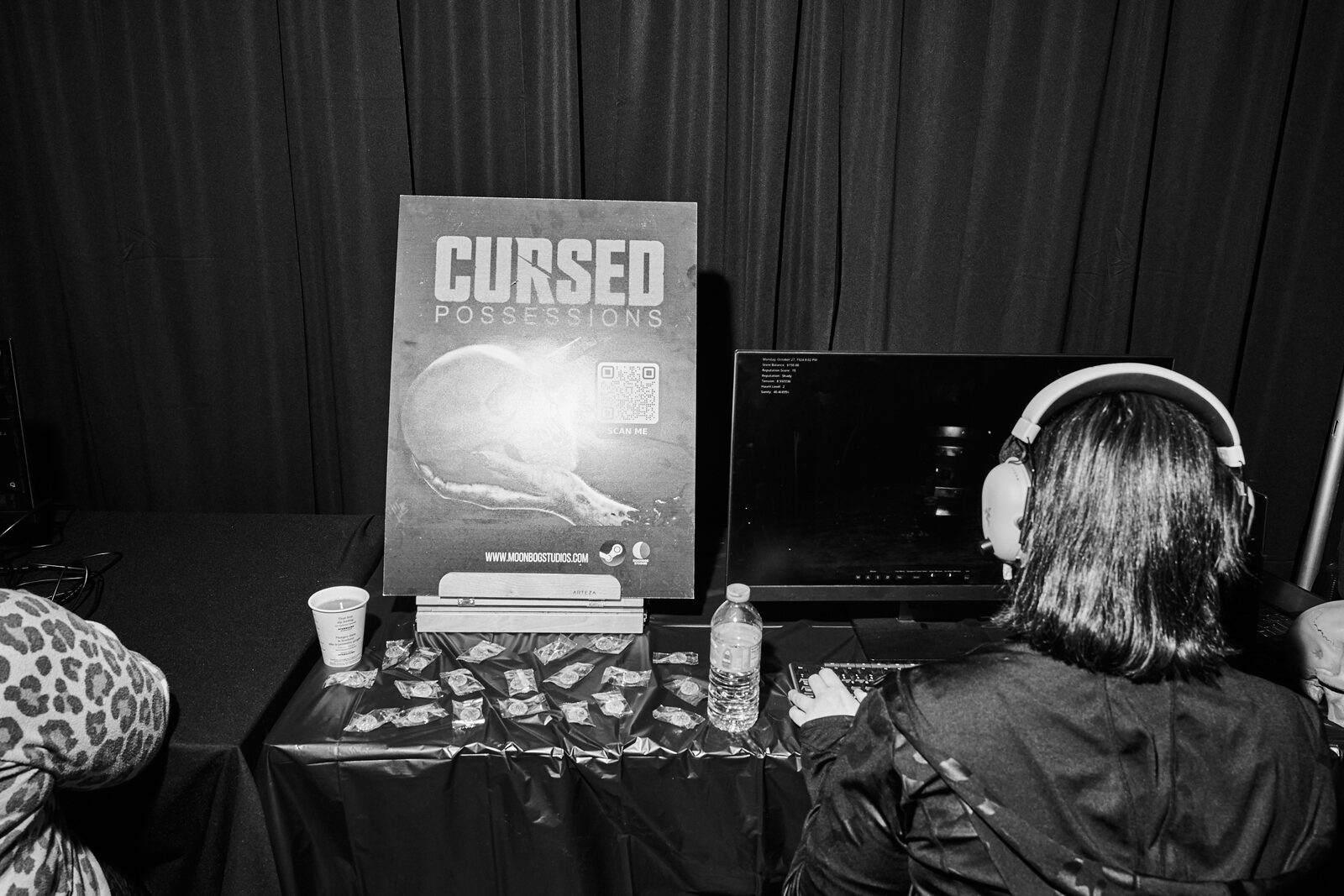 A showcase booth for Cursed Possessions — pull-up banner beside a monitor, a player in headphones at the keyboard.