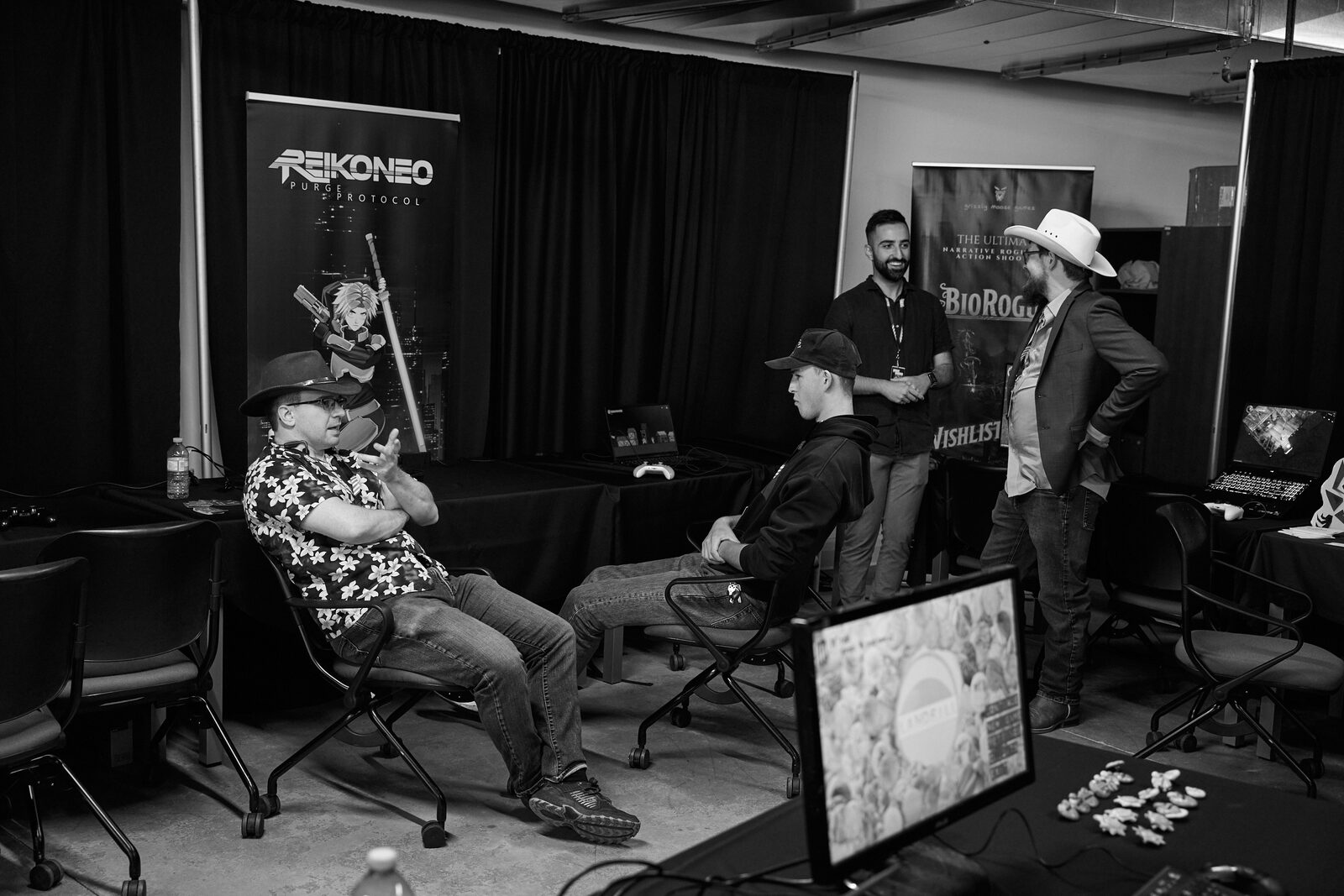 Four attendees in cowboy hats and caps talking between showcase stations, Reikoneo and BioRogue banners behind them.