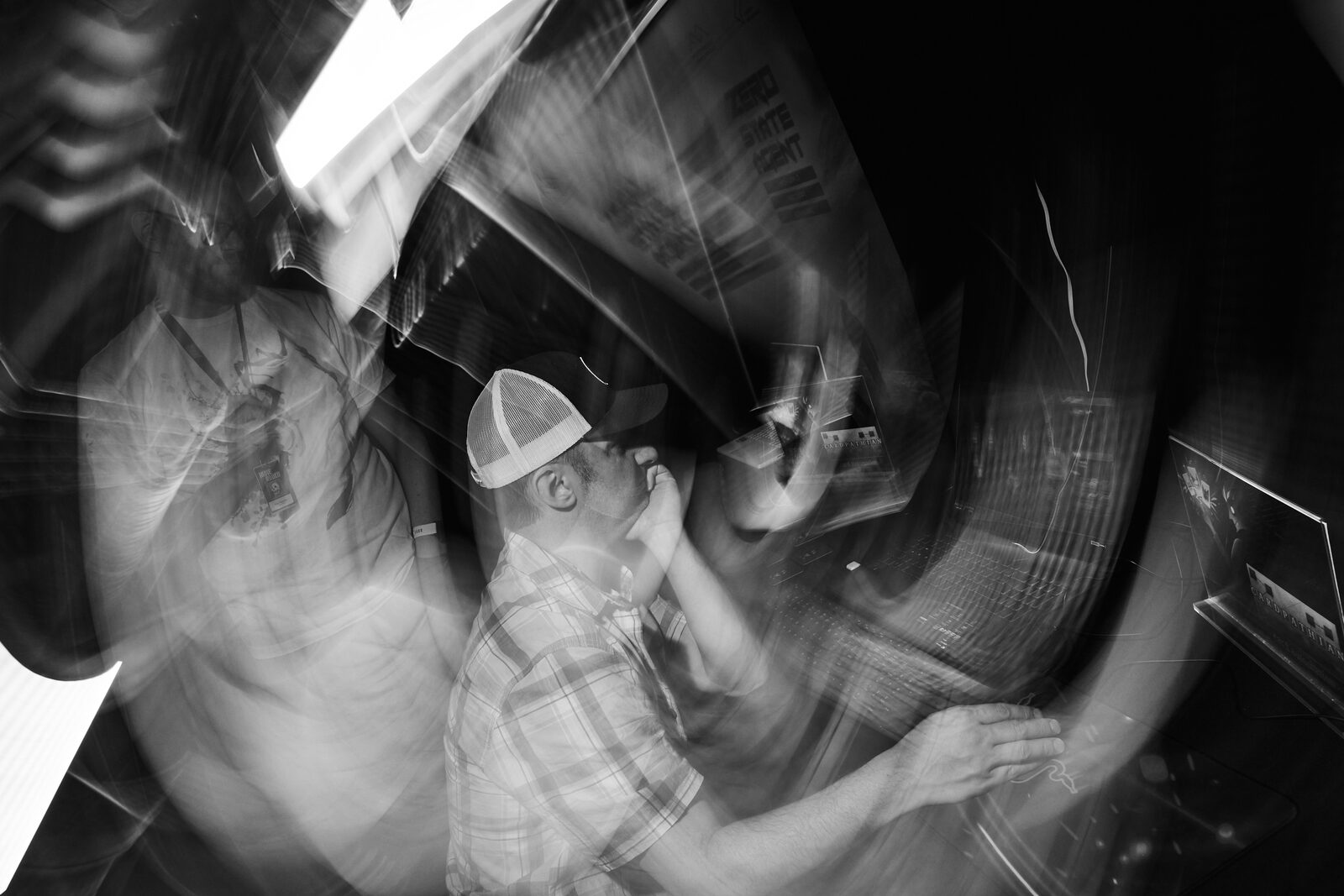 A spun long-exposure of the showcase floor: an attendee in a cap sharp at the centre while the booths smear into streaks around him.