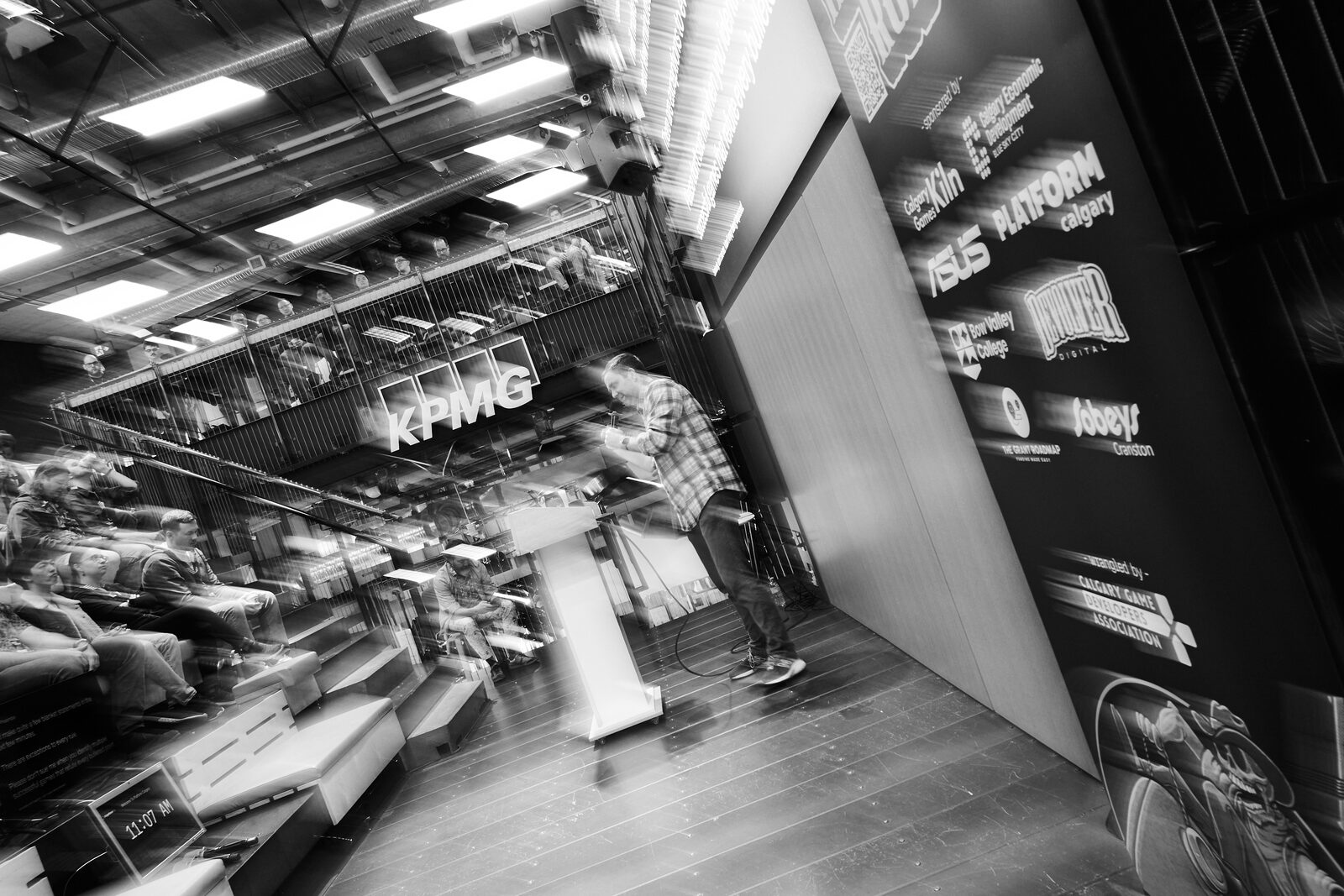 A motion-blurred wide of the atrium: the speaker at the podium held steady while the room swings past, sponsor wall to the right.