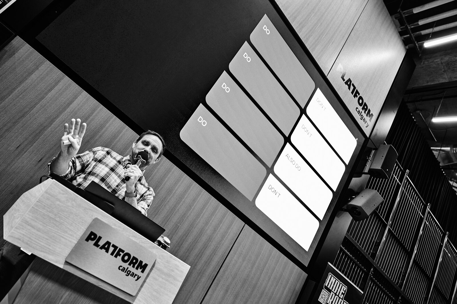 A speaker at the Platform Calgary podium with a hand up mid-point, their slide grid filling the screen behind.