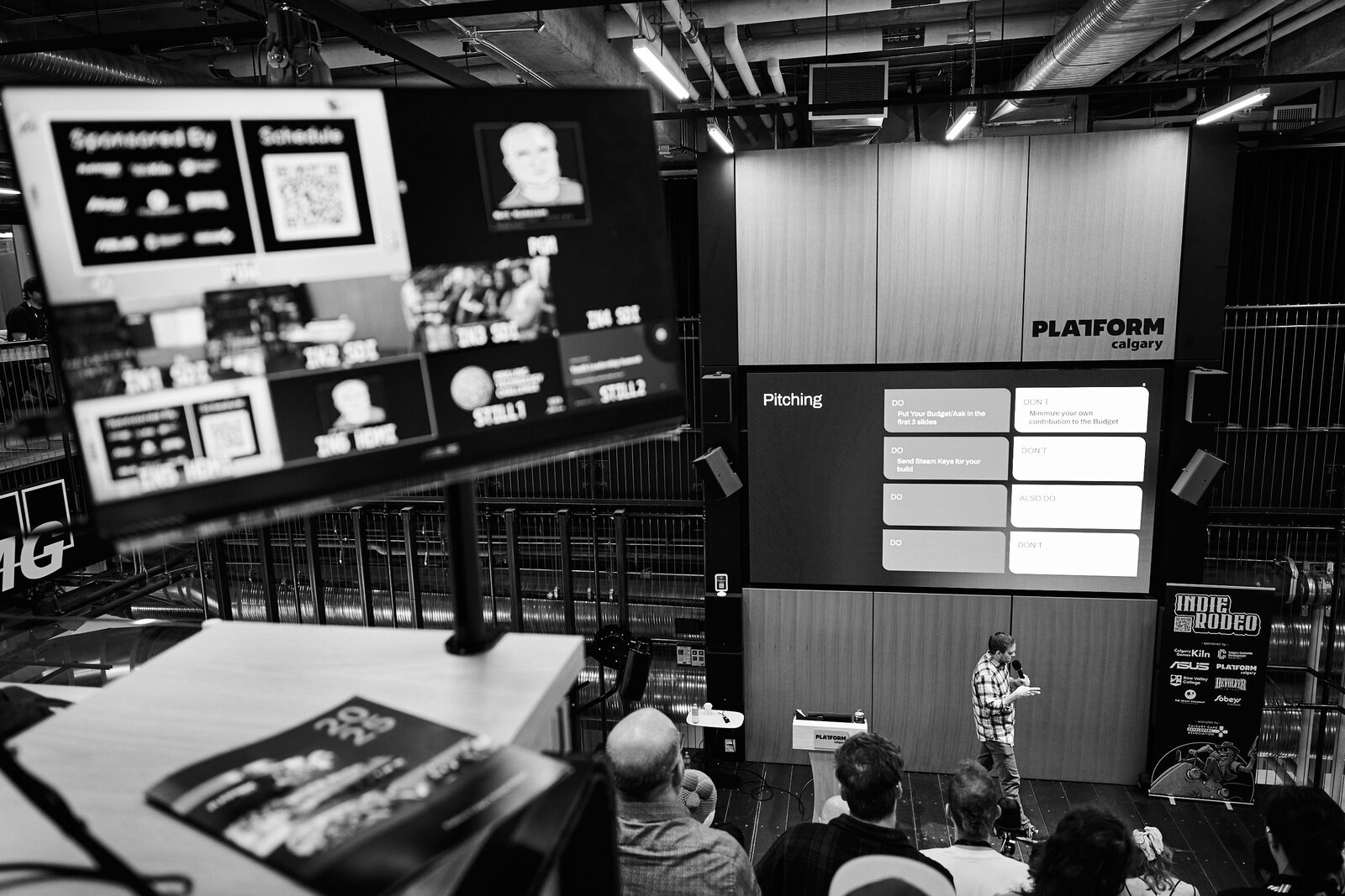 The atrium from the side: a "Pitching" slide on the main screen, the run-of-show and sponsor grid on a second monitor, the speaker small on stage.
