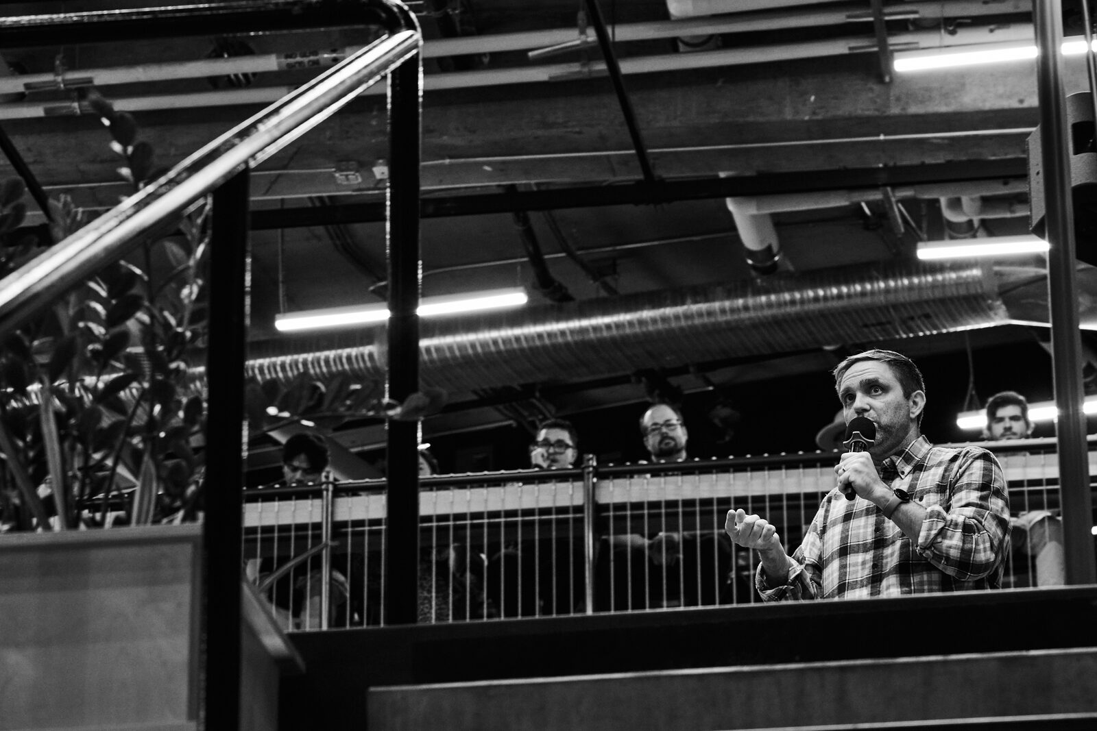 A speaker with a mic seen past the atrium railing, attendees watching from behind the mesh barrier.
