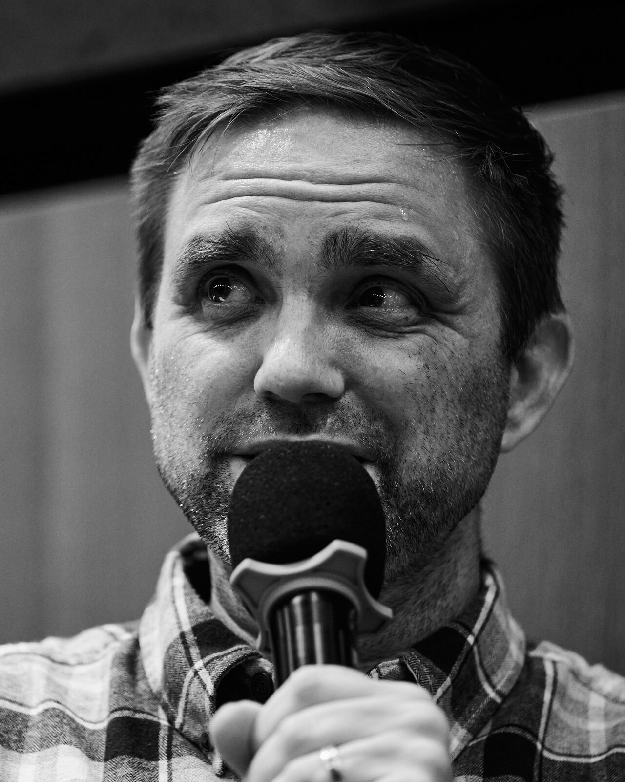 Tight portrait of a speaker in a plaid shirt holding the mic close, caught mid-thought.