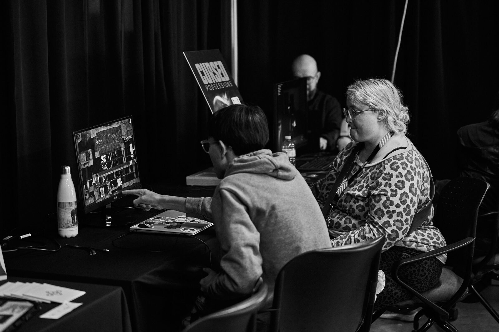 Two attendees at a showcase monitor, one pointing something out on screen, the Cursed Possessions banner beside them.