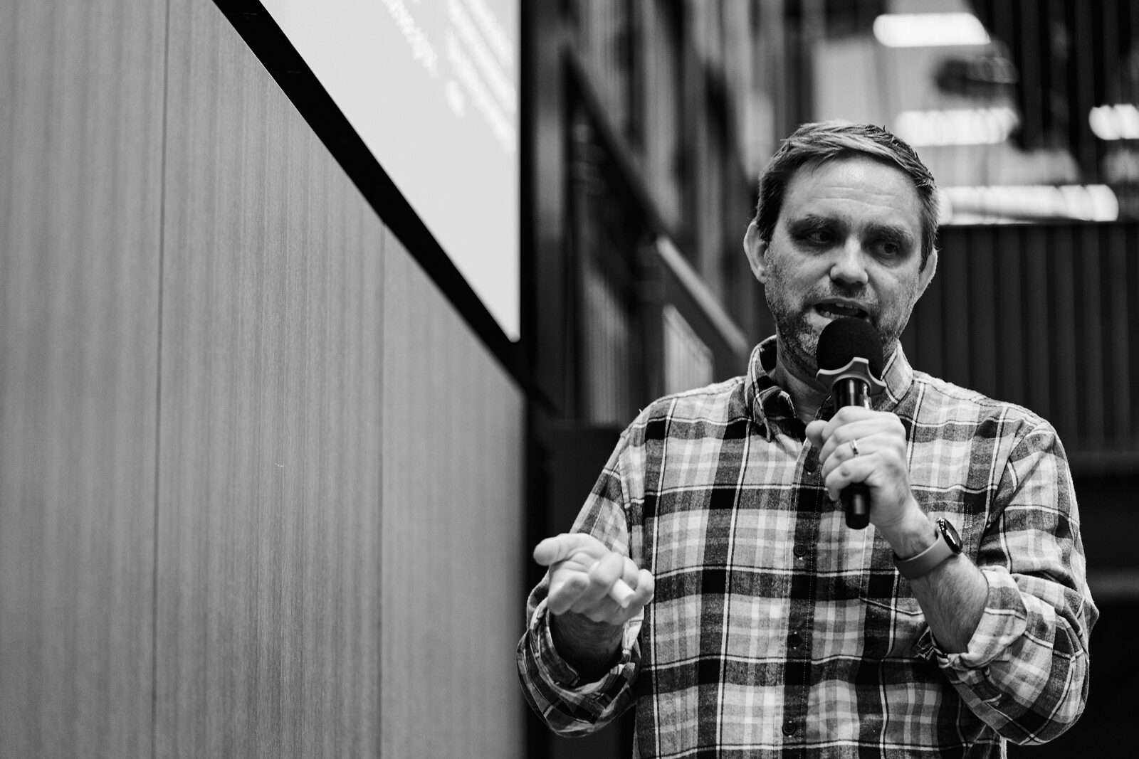 A speaker in a plaid shirt works the mic with his free hand raised, his slide glowing behind him.
