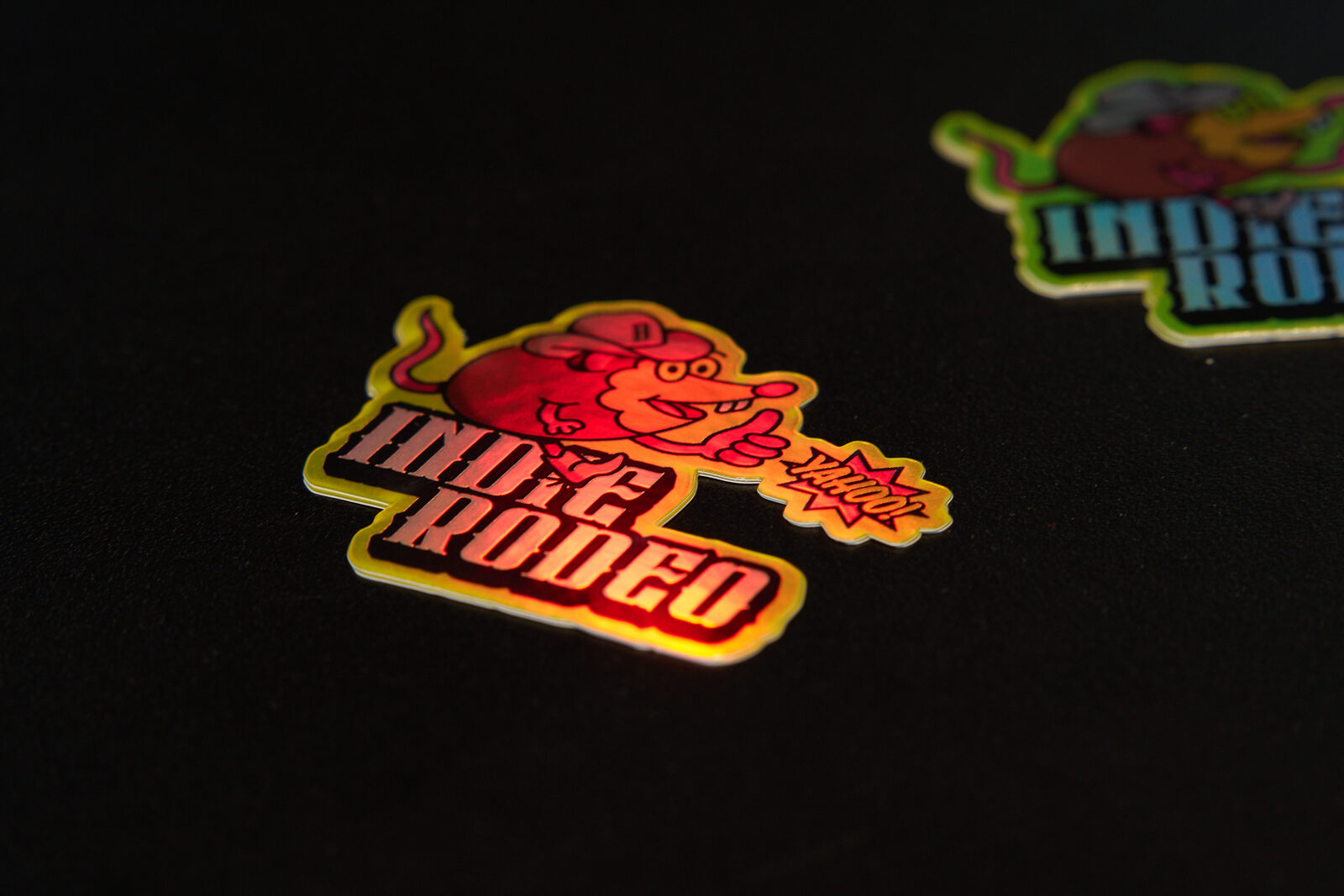 Two holographic Indie Rodeo stickers on a black surface, the rat mascot lit up in pink and orange.