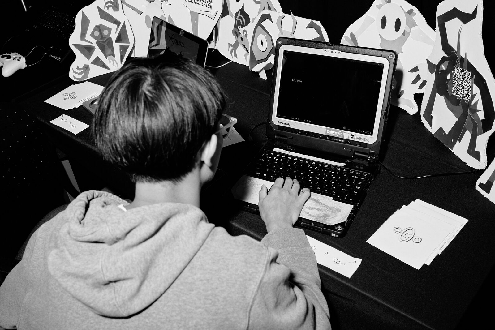 An attendee at a laptop on the showcase floor, ringed by paper standees and cut-out character art.