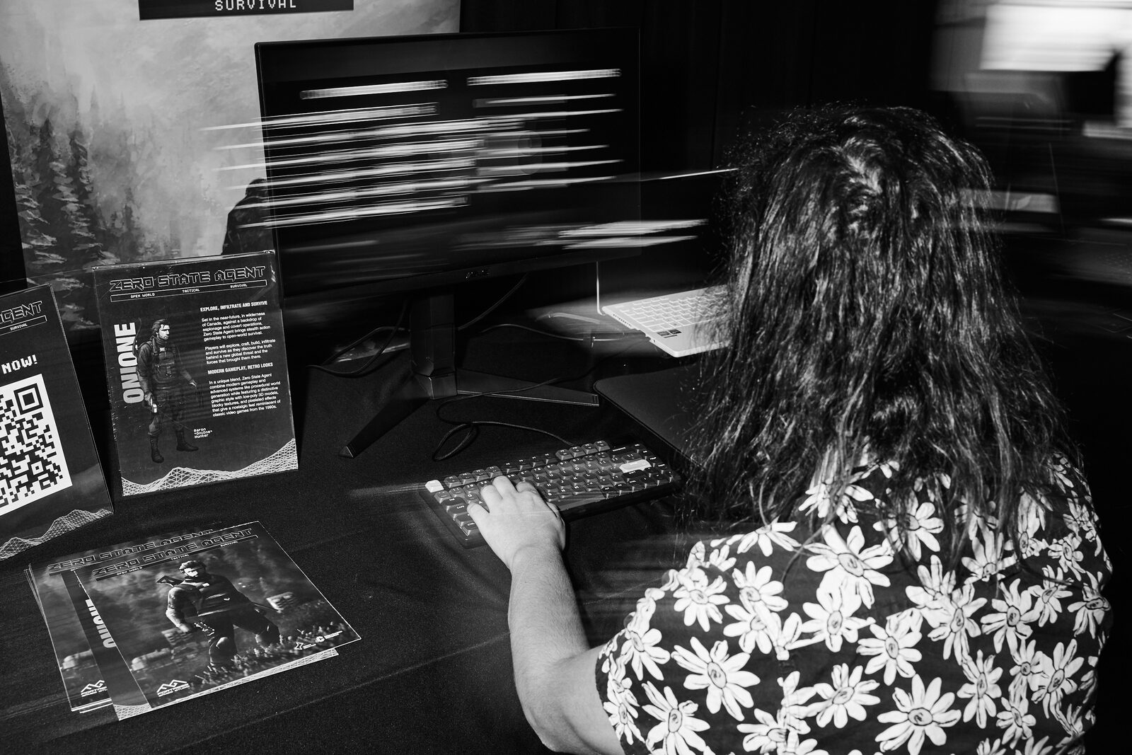 An attendee plays a demo at a showcase station, the monitor blurred with motion, flyers and a QR card on the table.