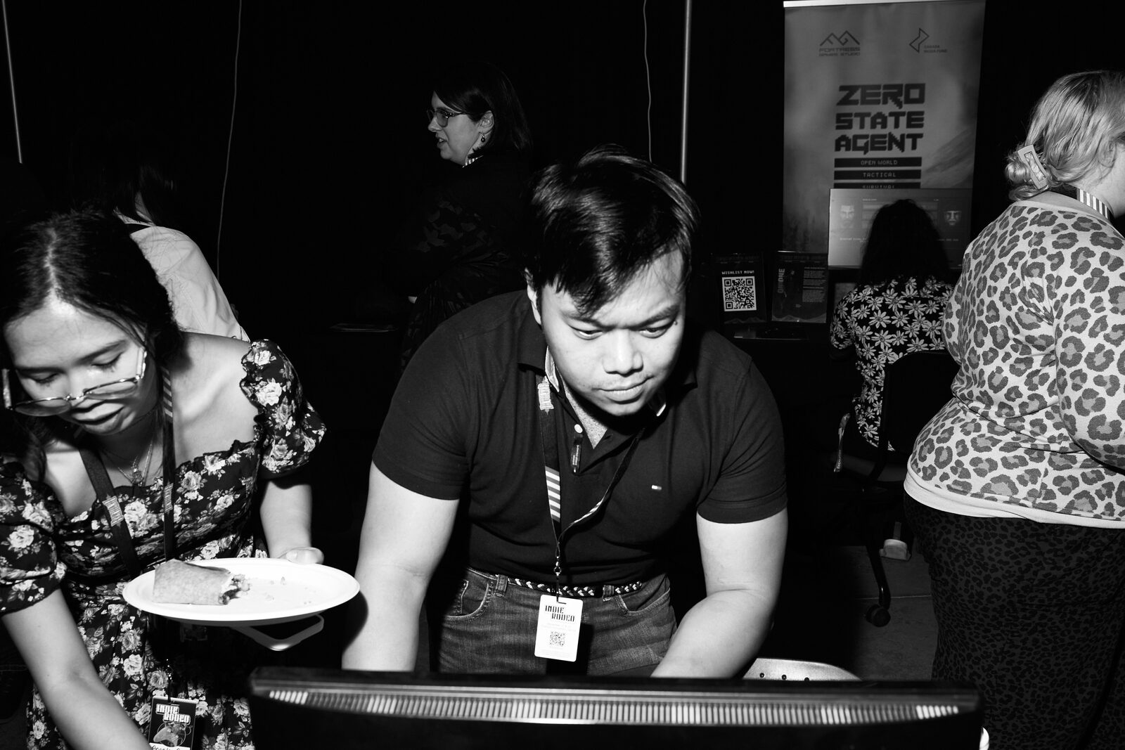 An attendee carries a plate of food past the showcase monitors while another leans in to play.