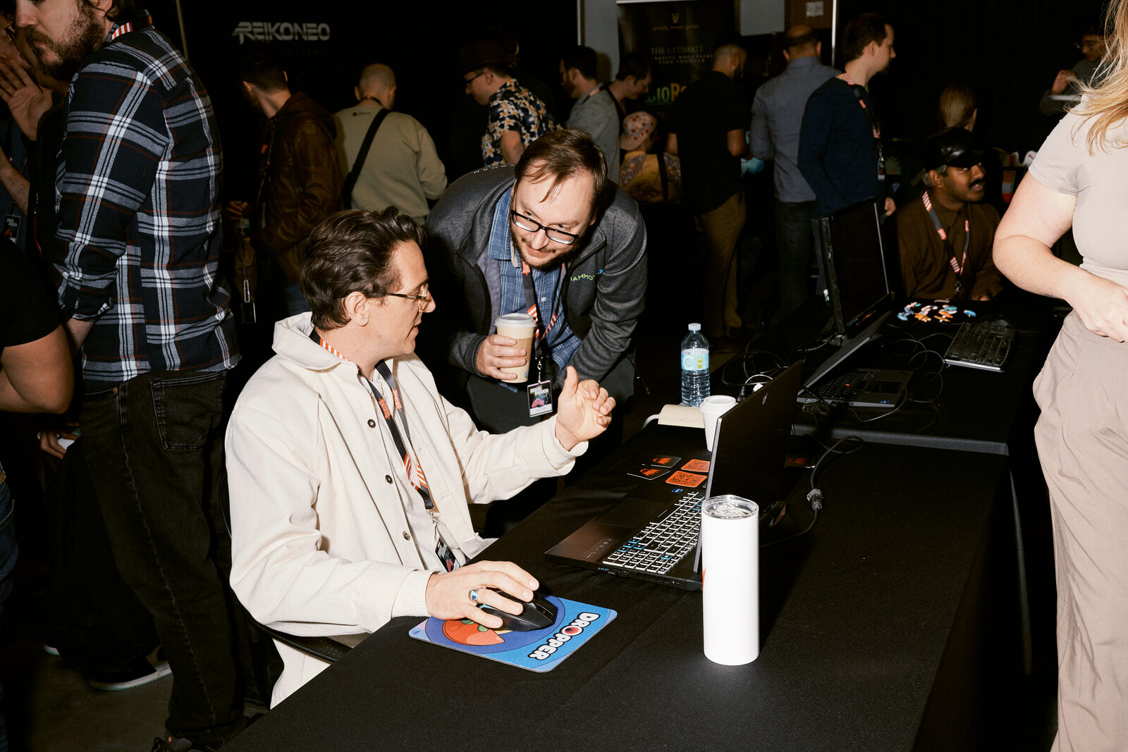 Two attendees at a showcase station on the crowded floor, one at the mouse, the other leaning in with a drink.