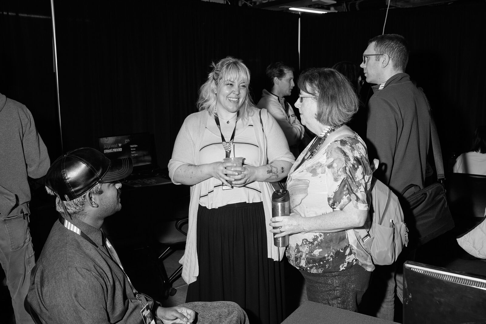 Attendees standing with drinks mid-conversation on the showcase floor, a seated attendee in a cowboy hat in the foreground.