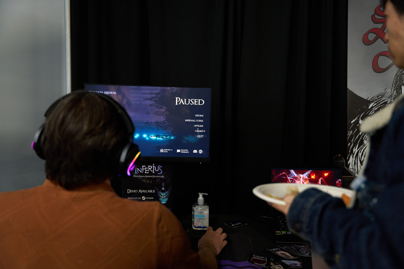 A player in headphones at the INFERIUS station, the game paused on screen, another attendee waiting beside him with a plate.