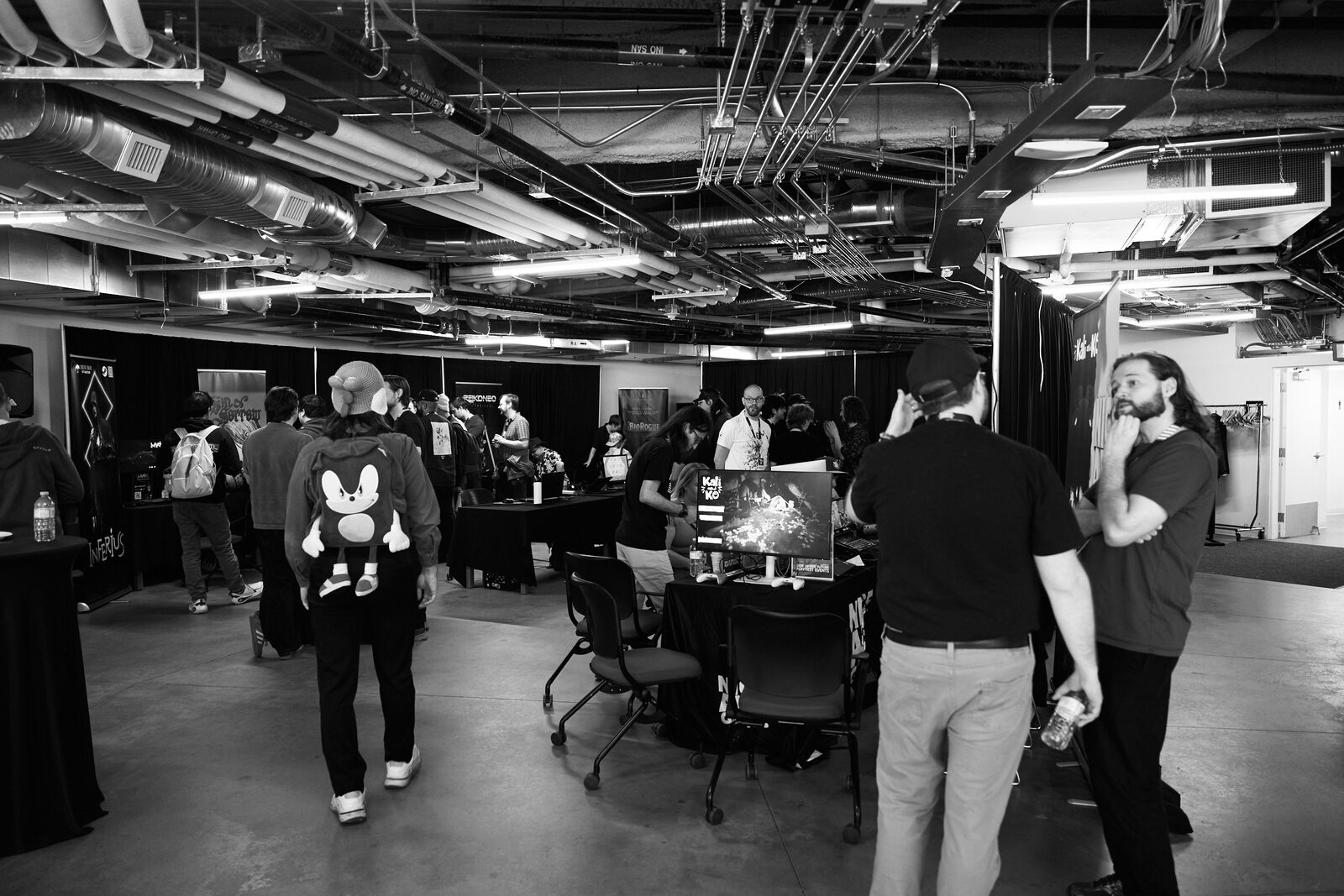 Wide of the showcase room under its exposed ceiling: attendees moving between stations, one with a Sonic backpack.