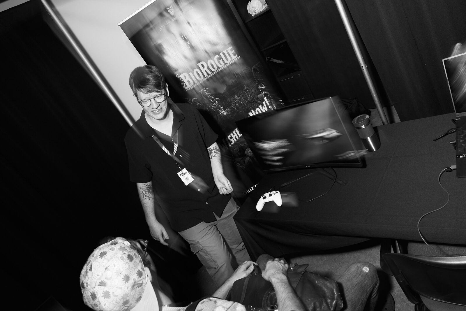 A developer stands grinning beside the BioRogue banner while a seated attendee plays his demo, controller in hand.