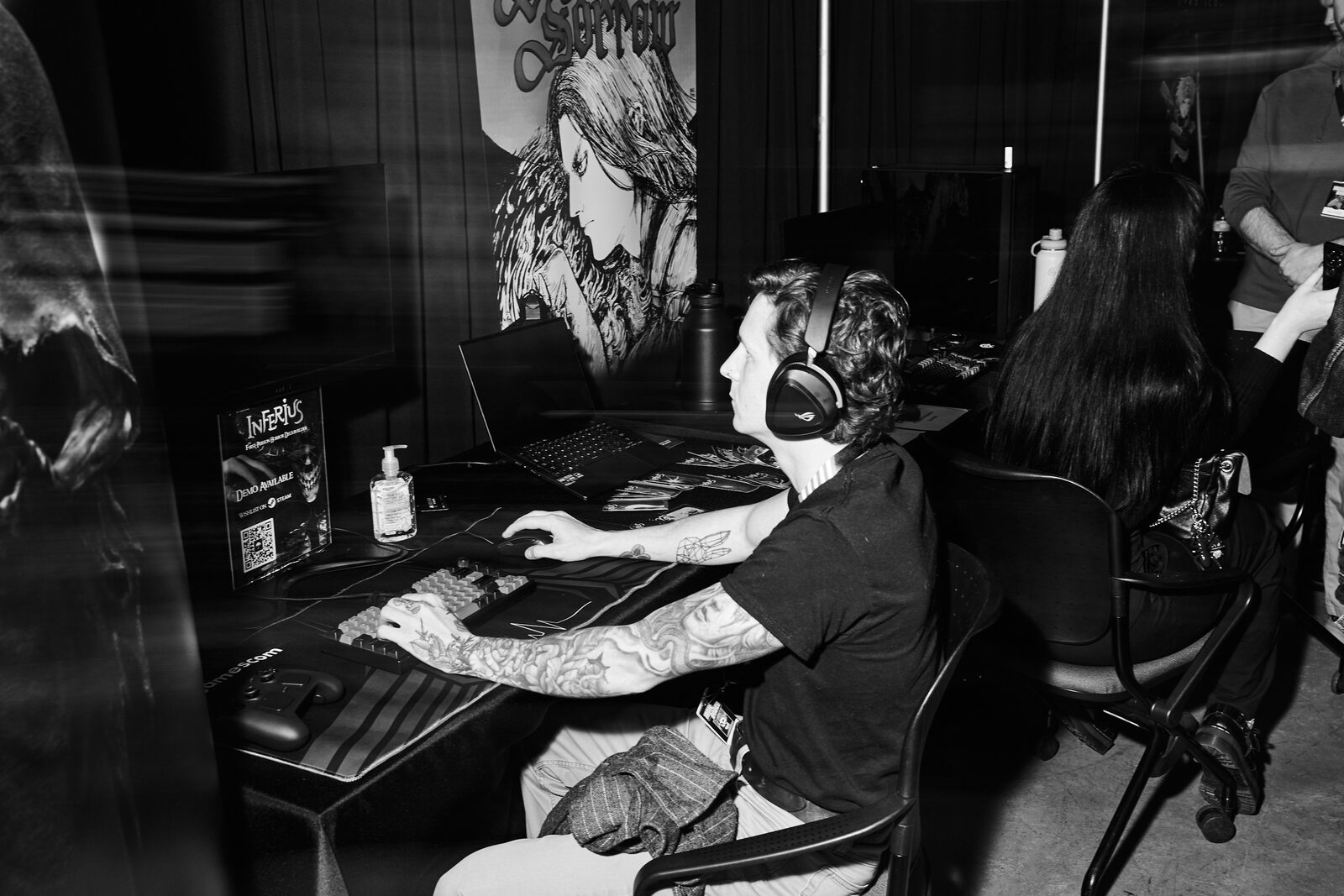 A player in headphones at a keyboard, tattooed arms on the desk, a large illustrated poster over the station.